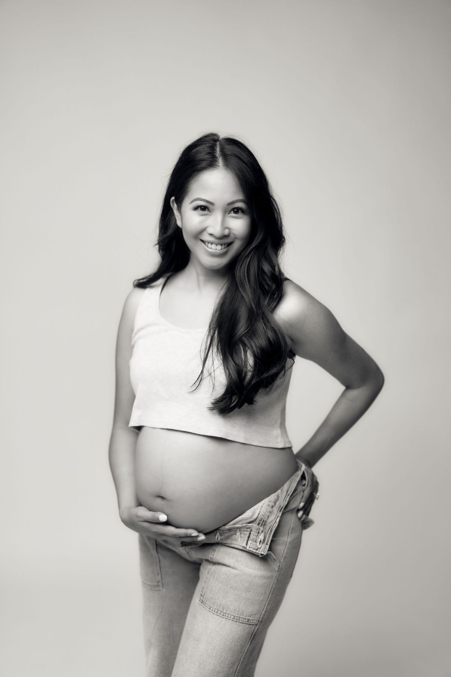 Black and white maternity portrait showing an expectant mother in a crop top and jeans against a neutral backdrop.