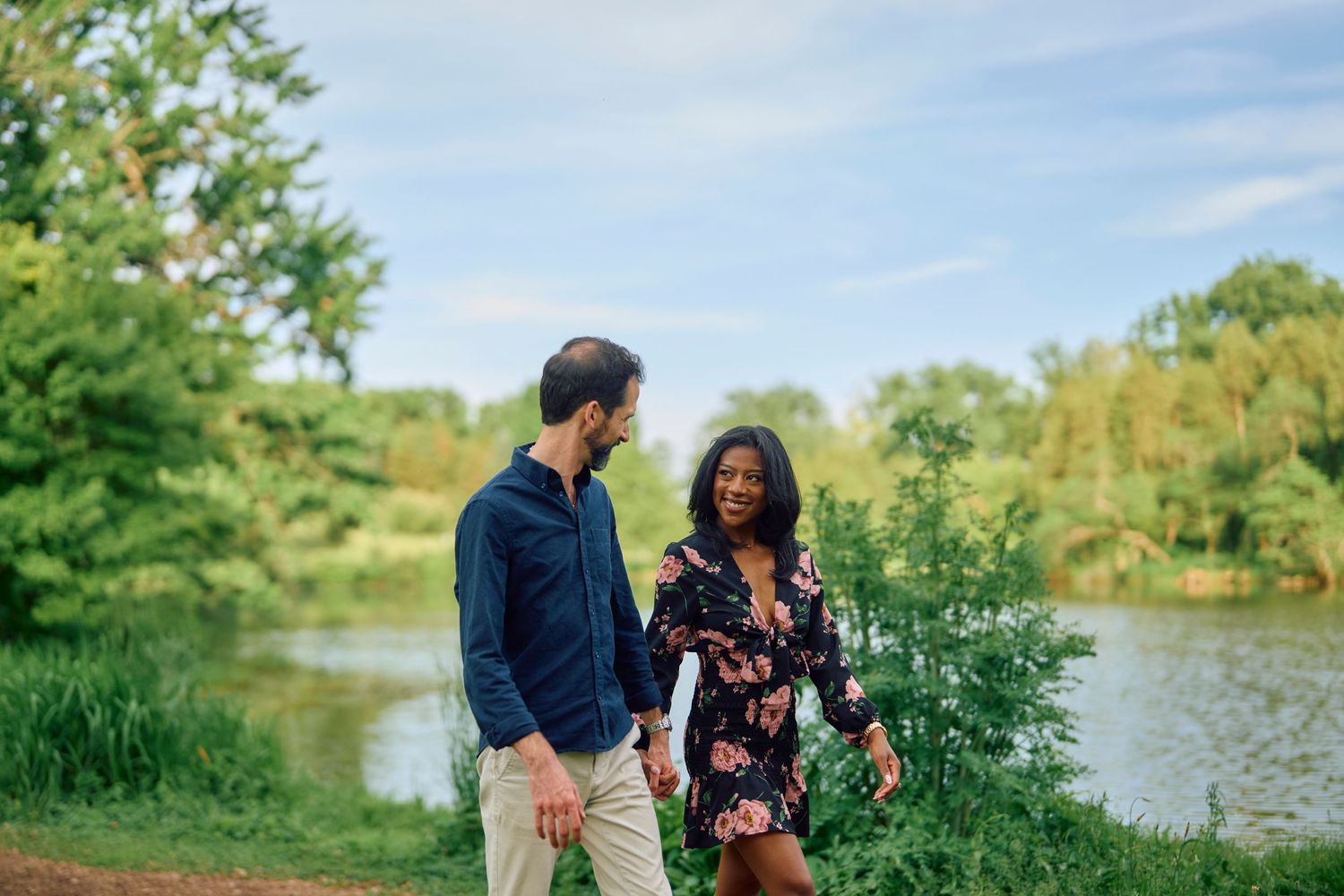 A couple enjoys a romantic moment while walking along a scenic pond surrounded by lush greenery on a sunny day.