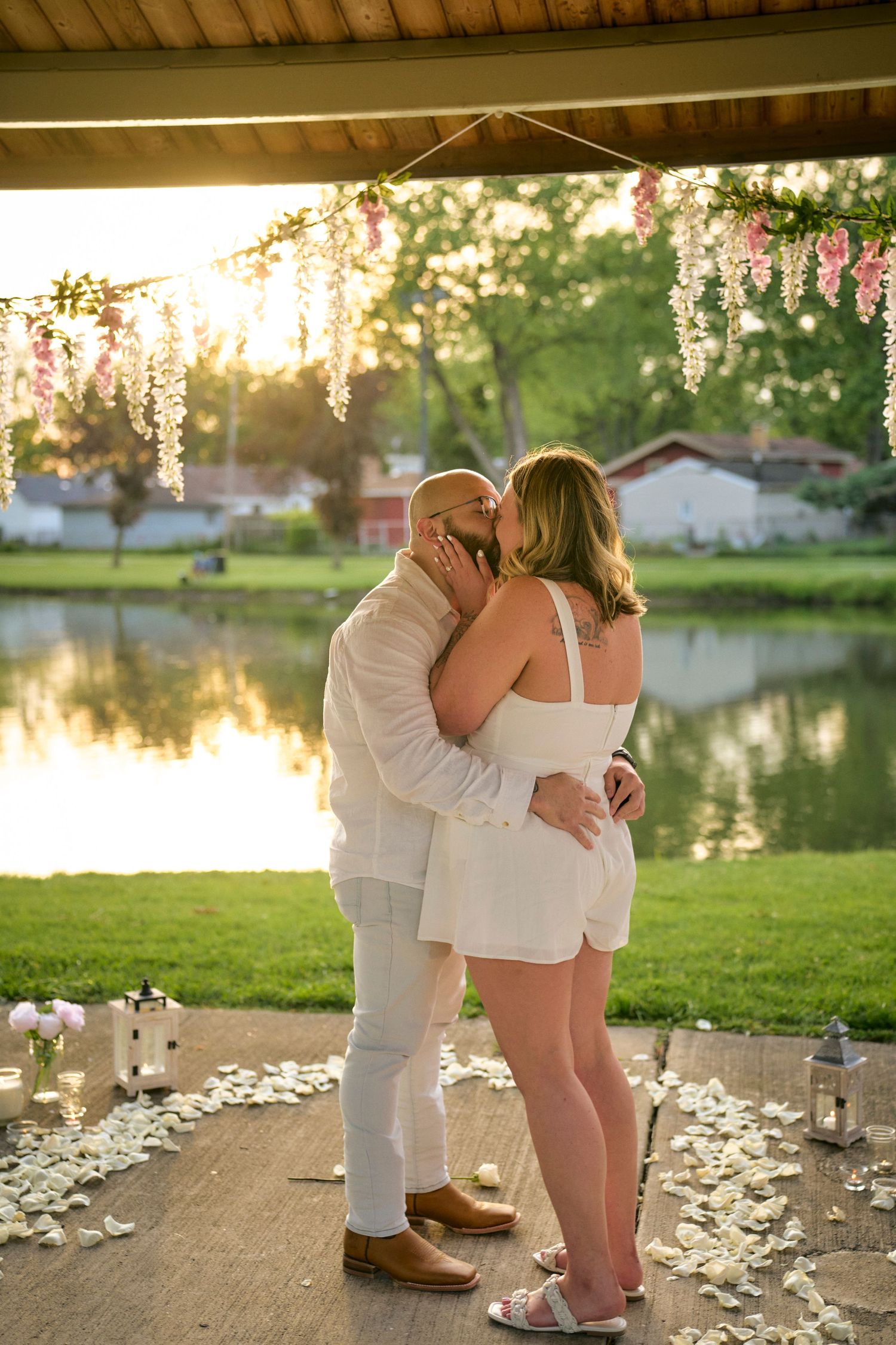 A romantic sunset engagement photo by a lake with hanging wisteria flowers and lantern-lined path.