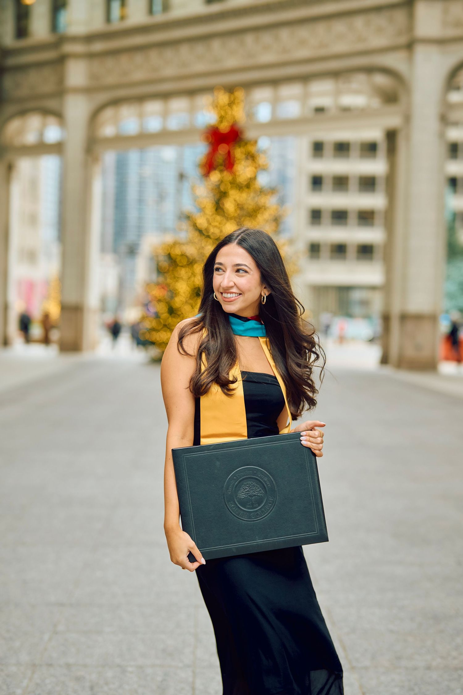 Stylish woman in a black dress and yellow scarf holds a shopping bag on a city street with a golden Christmas tree behind.