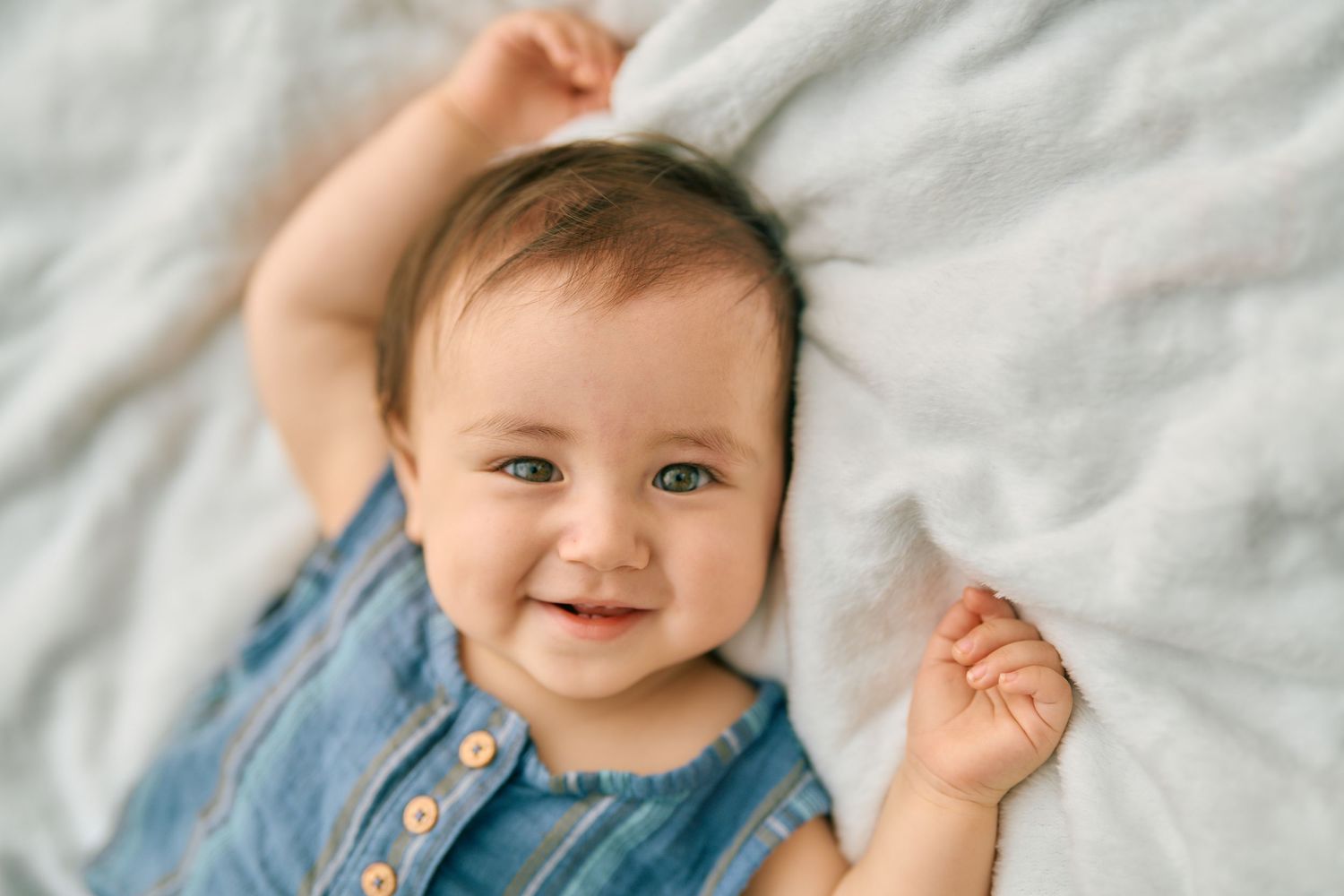 Happy baby in denim outfit laying on white bedding with arms raised joyfully.