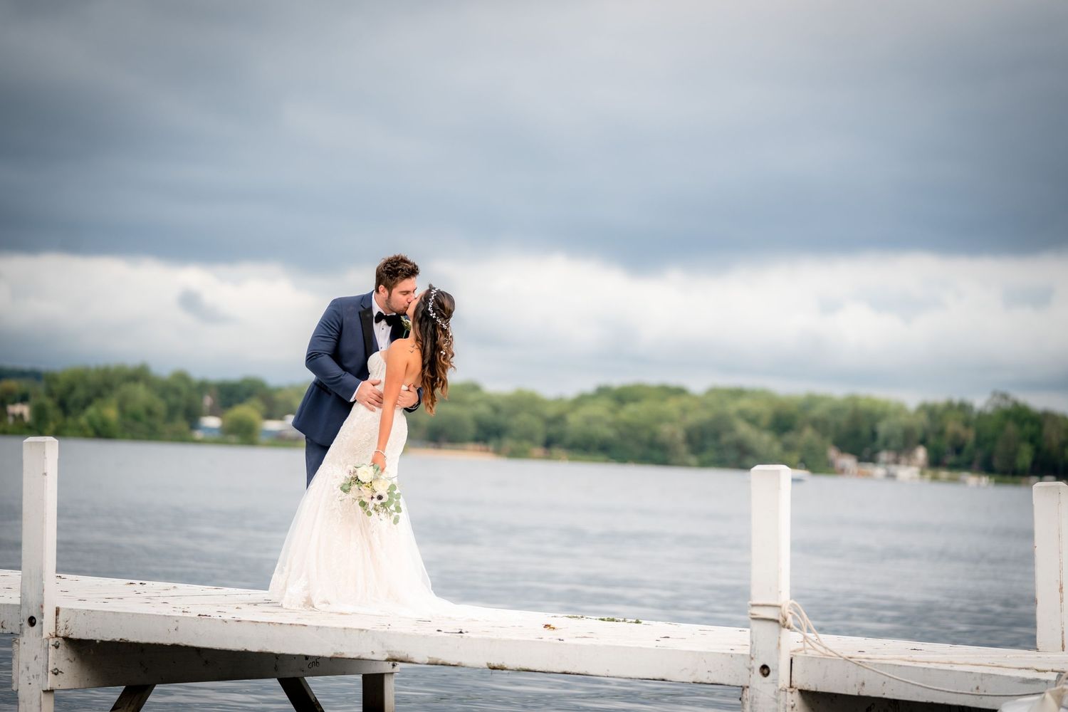 A romantic wedding couple shares an intimate moment on a wooden dock overlooking a serene lake under cloudy skies.
