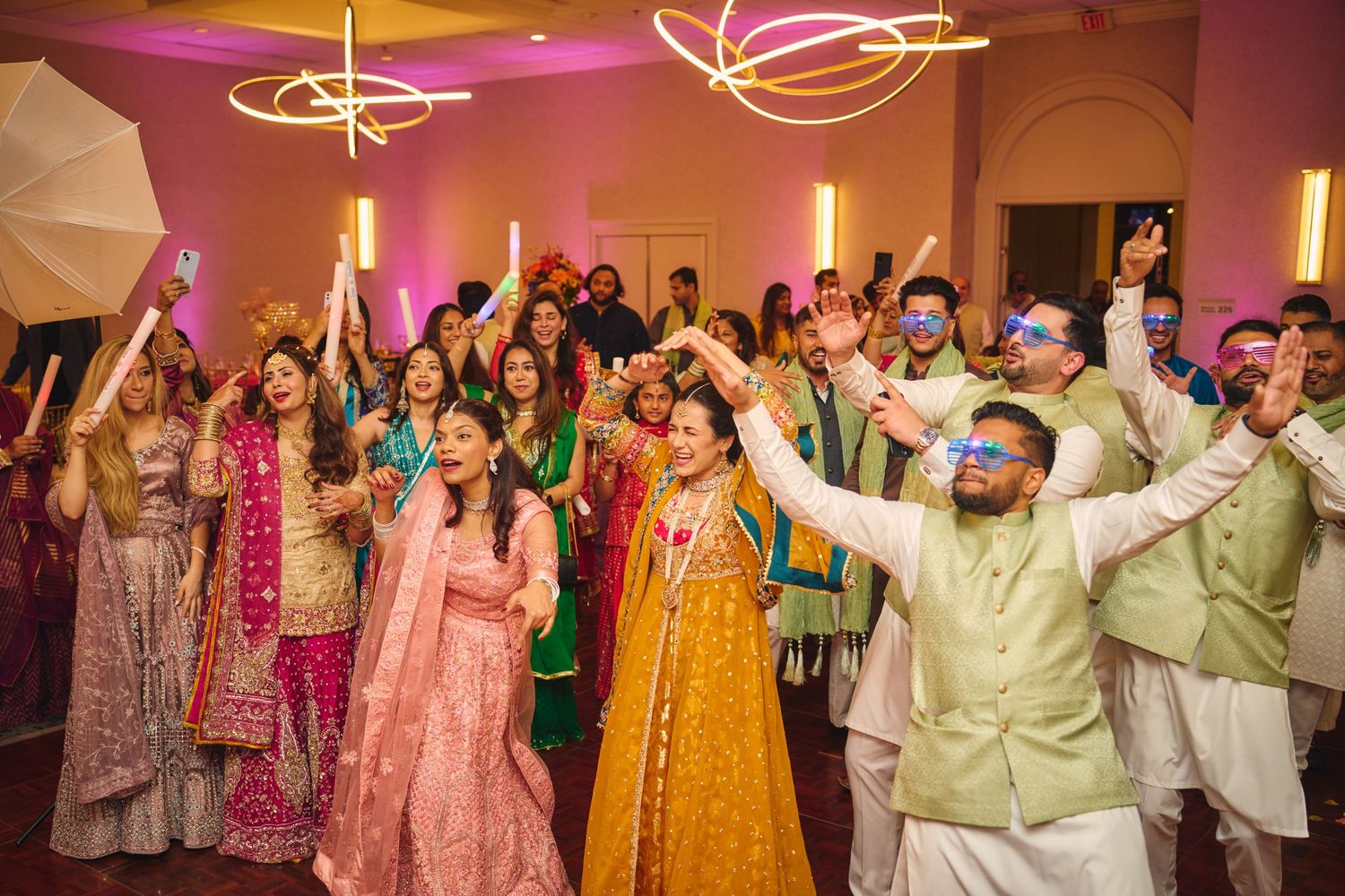 Guests dance and celebrate at a vibrant Indian wedding reception with pink and yellow lighting.