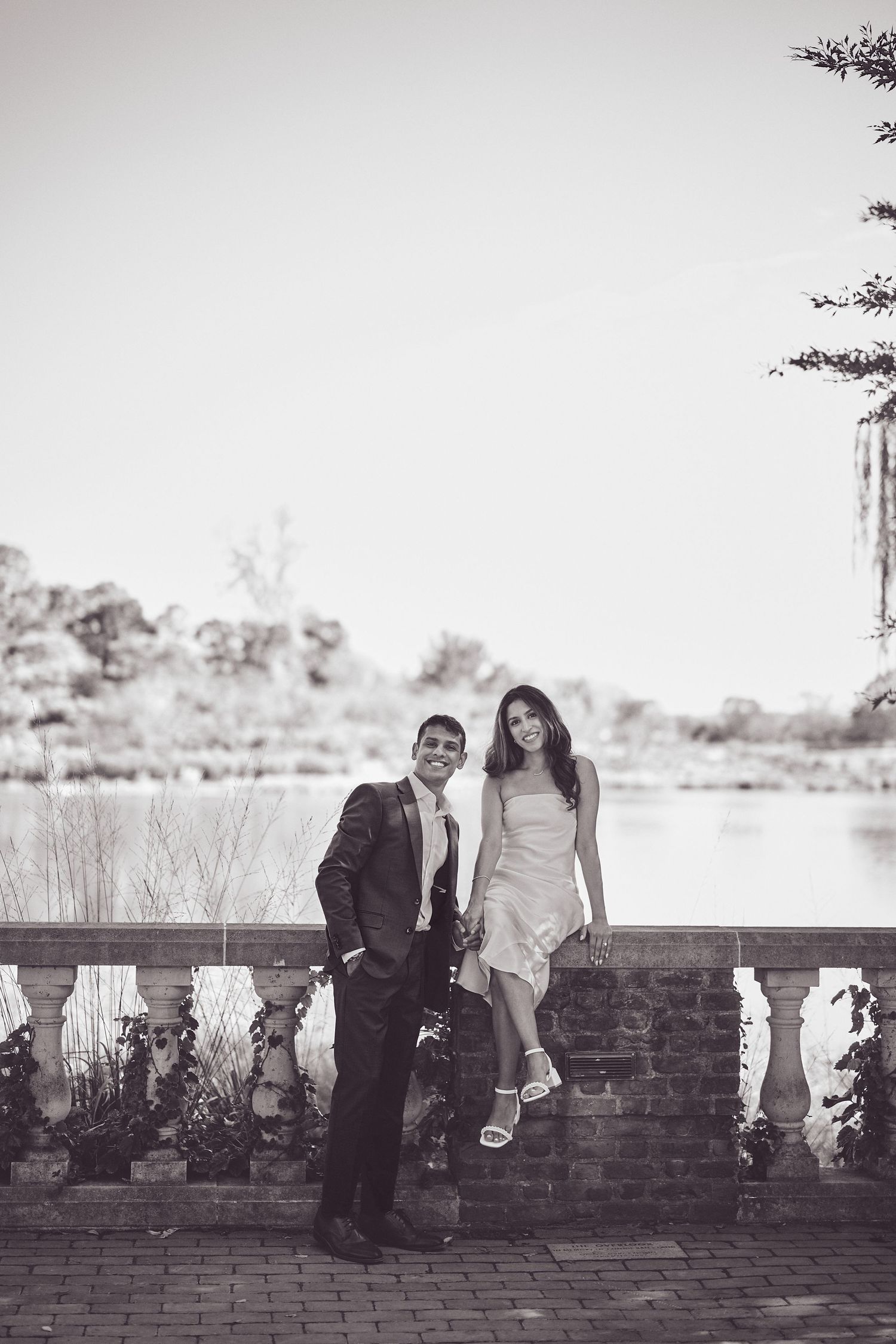A romantic couple poses together on an ornate stone balustrade by a lake in black and white photography.