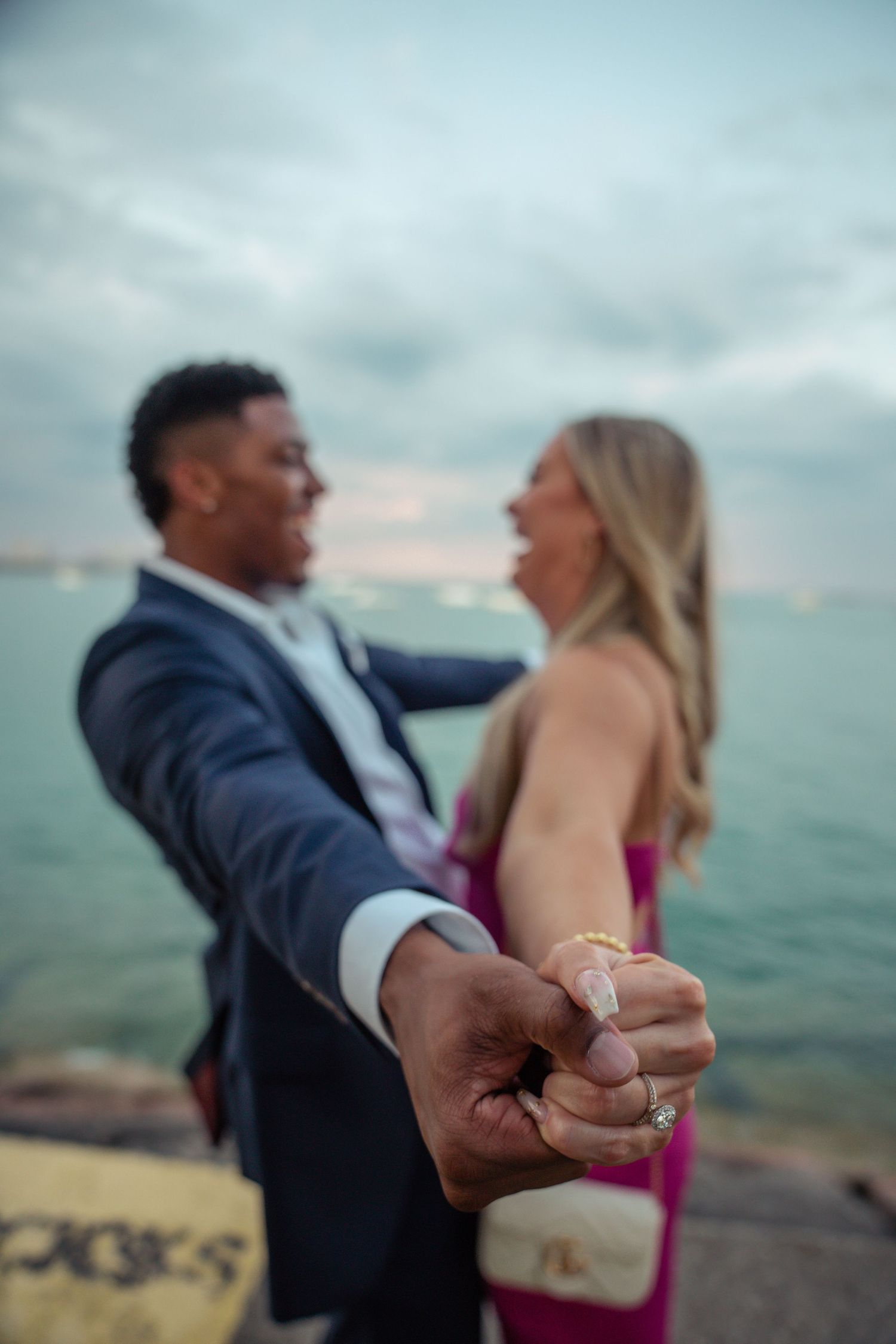 A couple shares an intimate engagement moment by the ocean as a close-up showcases their hands with a ring.