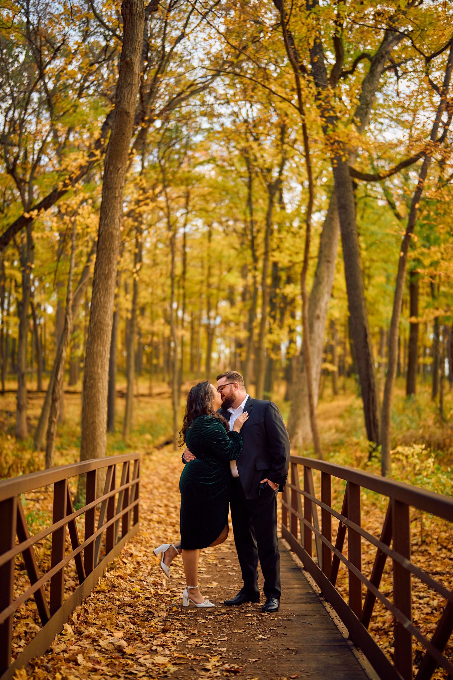 A romantic couple embraces on a rustic wooden bridge surrounded by golden autumn foliage in a peaceful forest setting.