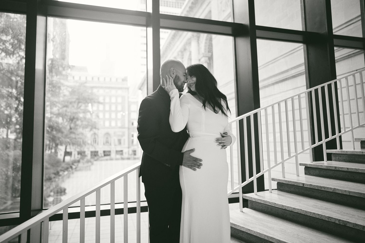 A couple in wedding attire share an intimate moment on a staircase with large windows and natural light.