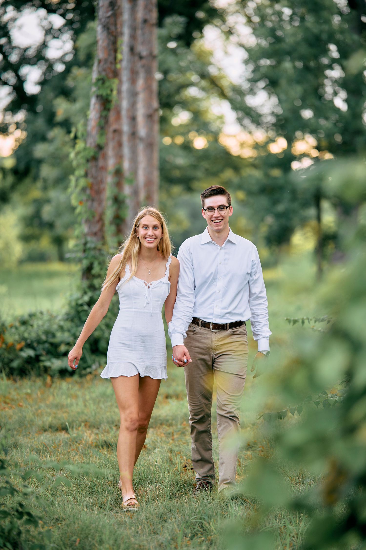 A couple walks hand in hand through a sunlit wooded area, wearing summer attire on a warm evening.