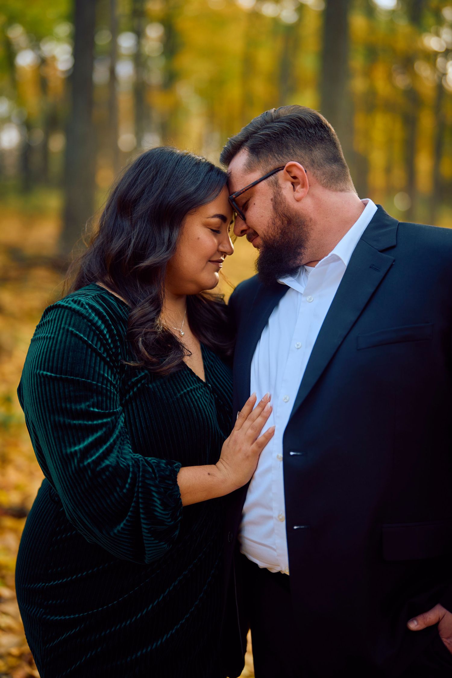 A romantic couple shares an intimate moment in a forest with golden autumn foliage in the background.