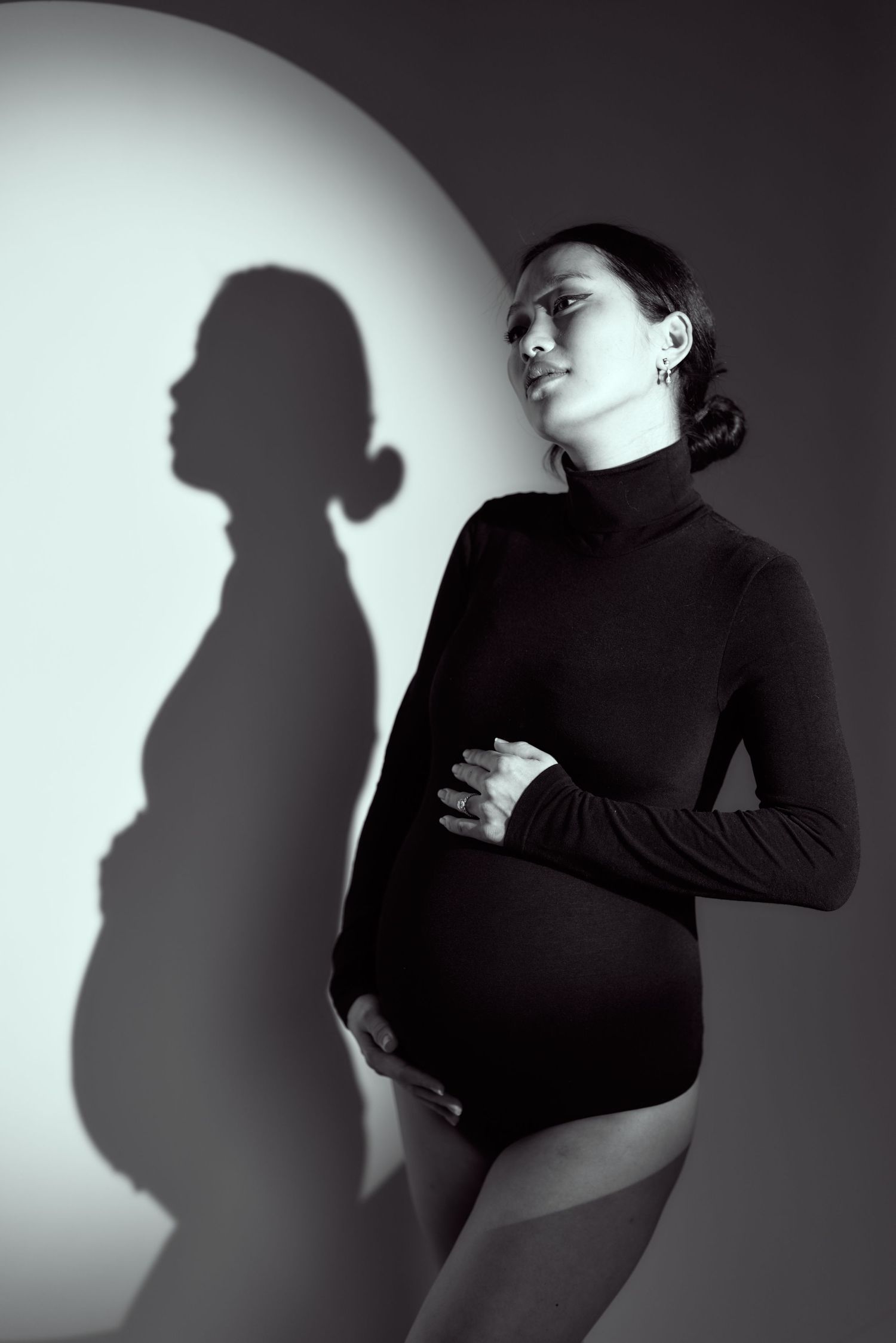 Artistic black and white maternity photo showing silhouette of pregnant woman in turtleneck against light wall.
