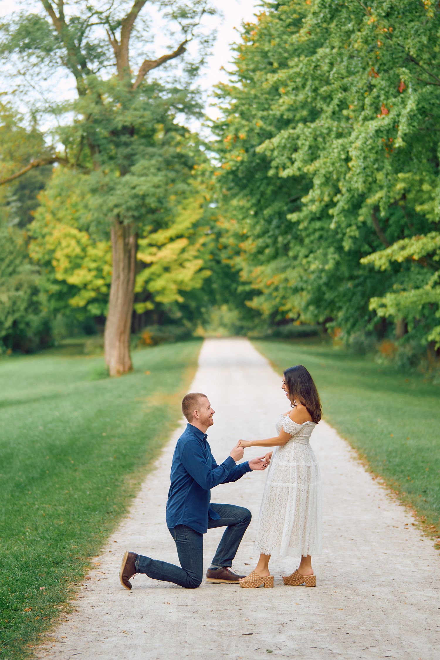 A romantic proposal scene on a tree-lined gravel path in a lush green park setting on a sunny day.