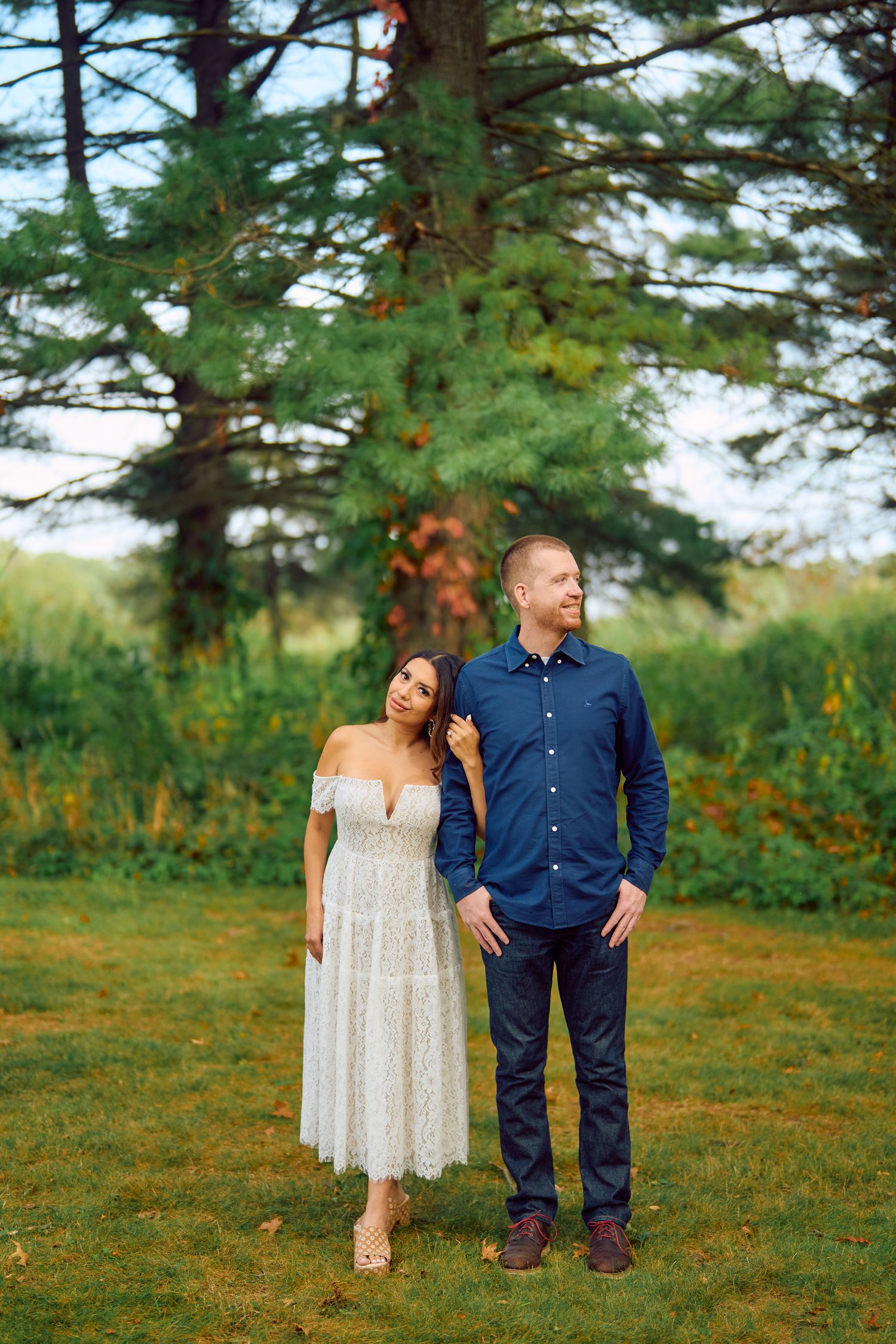 A couple stands together under evergreen trees on a grassy lawn, with the person in lace dress leaning on their partner.