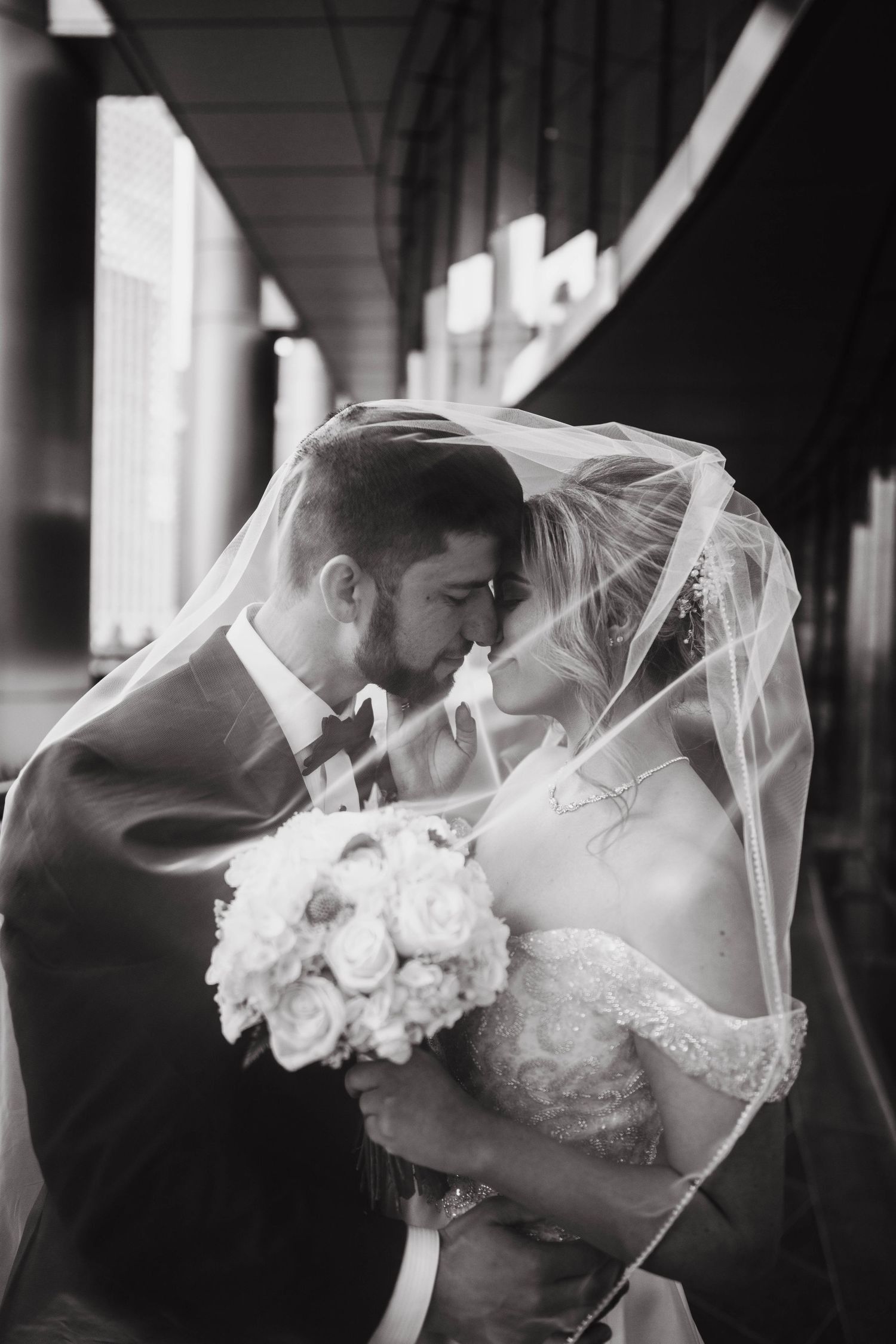 A romantic black and white wedding photo with the couple embracing under a flowing veil, holding a white rose bouquet.
