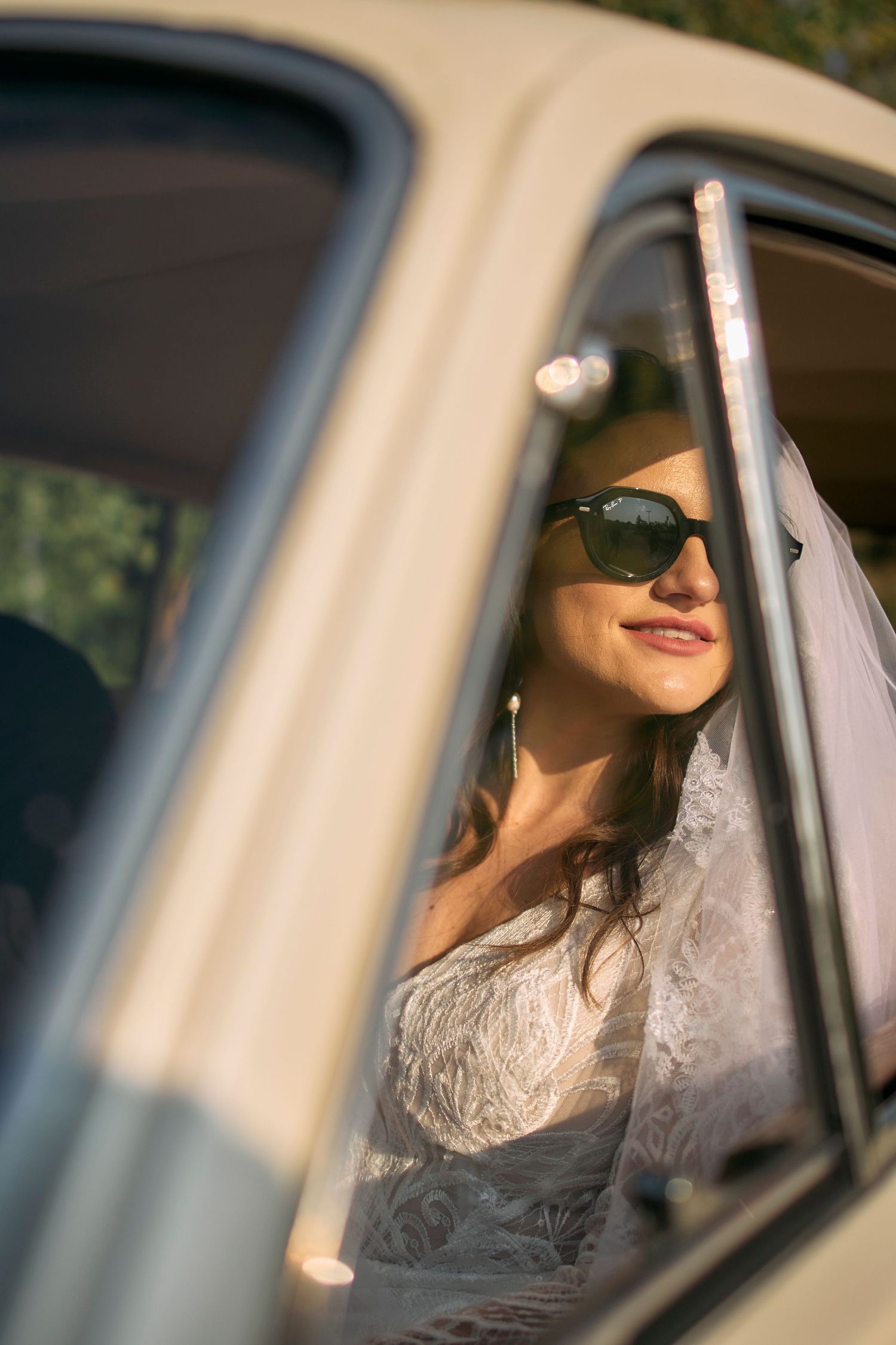 Bride wearing sunglasses smiles from inside a classic vintage car window.