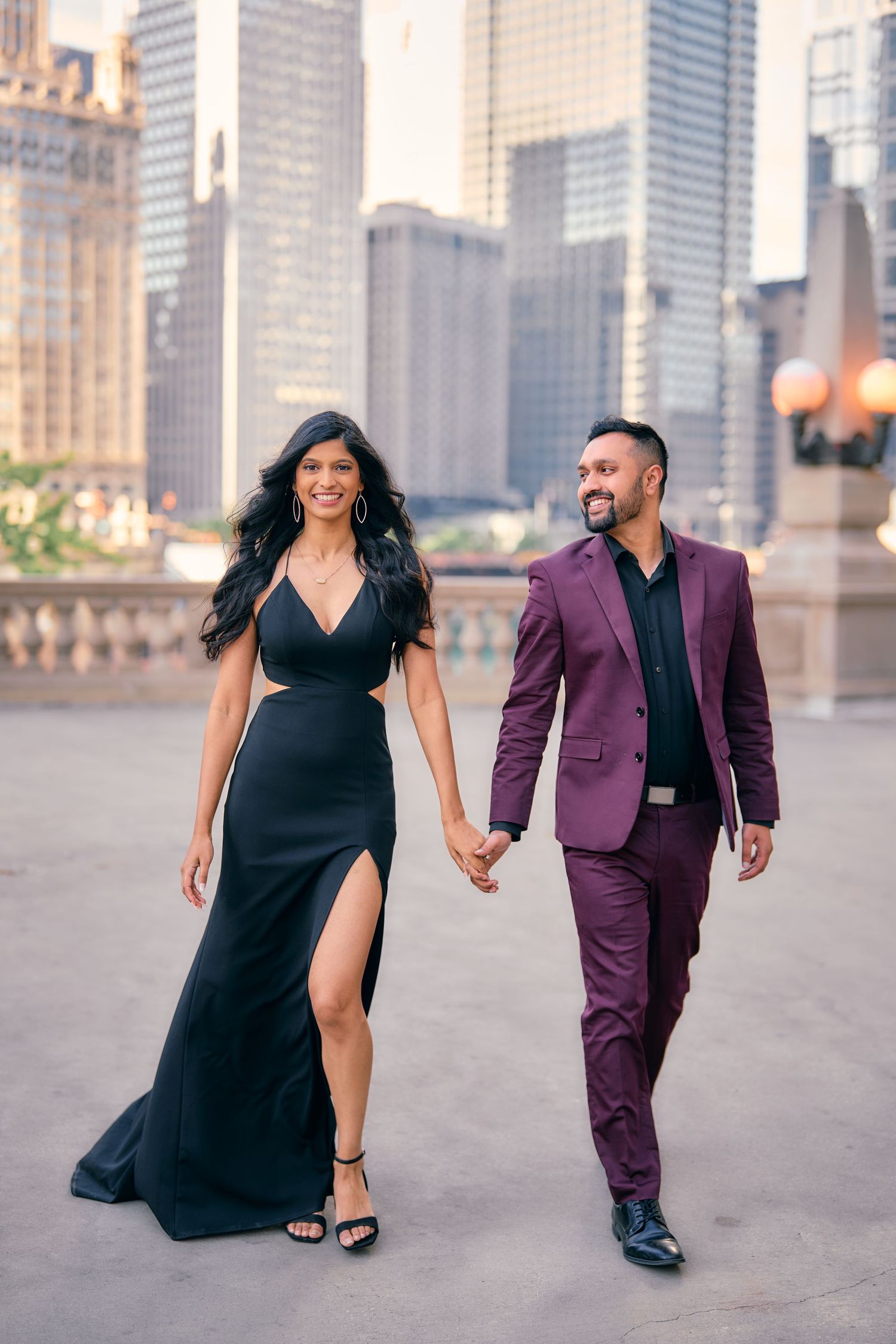 A stylish couple walks hand in hand in formal wear against a cityscape backdrop.