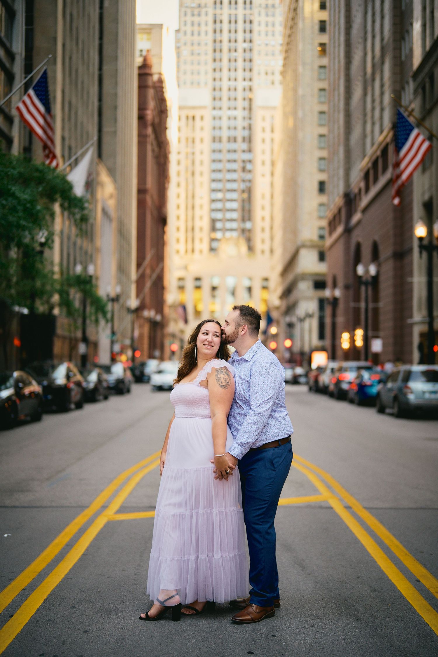 A romantic couple poses on a city street with American flags and skyscrapers lining the backdrop.