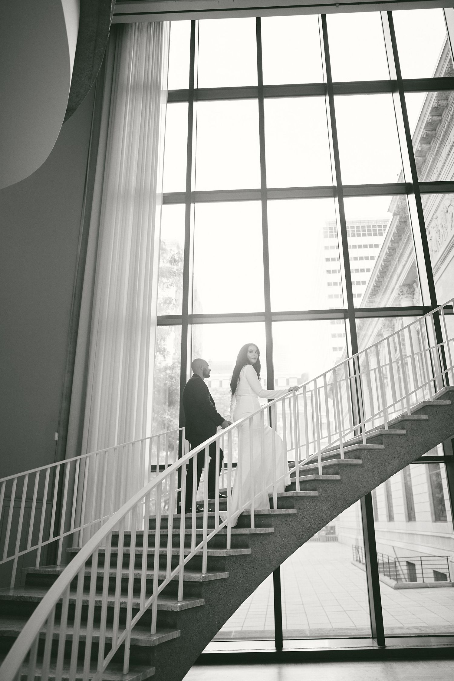 A couple ascends a modern white interior staircase beside large windows in a black and white photograph.