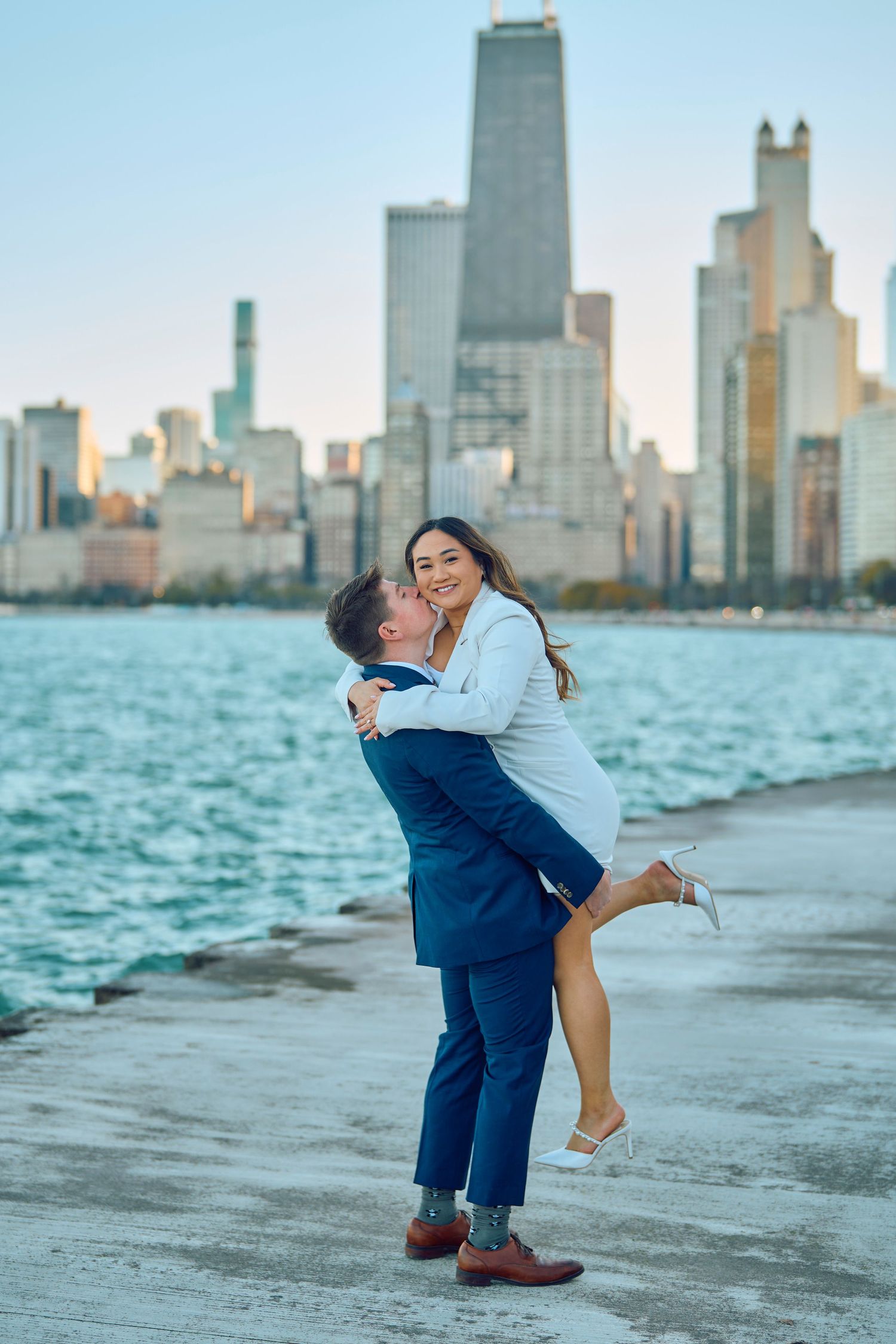 Couple embraces on lakefront path with Chicago skyline and Lake Michigan waters in background during daytime.