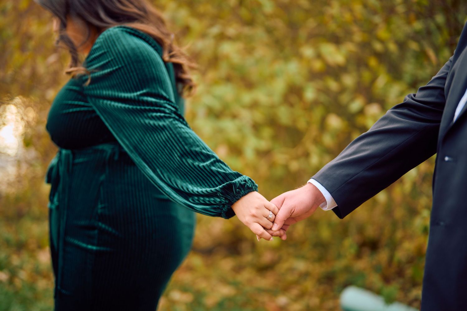 Two people hold hands against an autumn background with golden bokeh and fallen leaves.