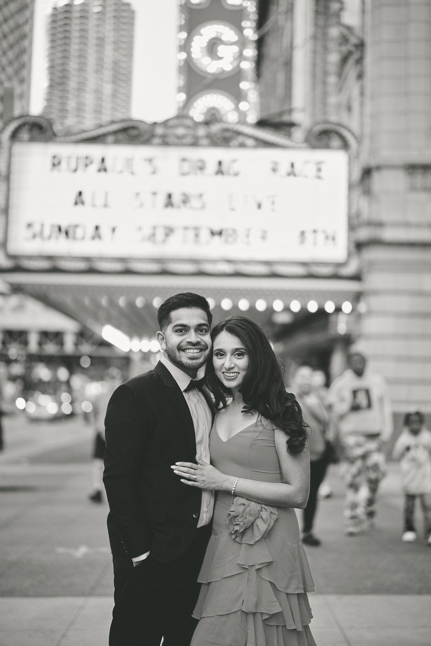A couple poses together outside a vintage theater marquee in black and white photography.