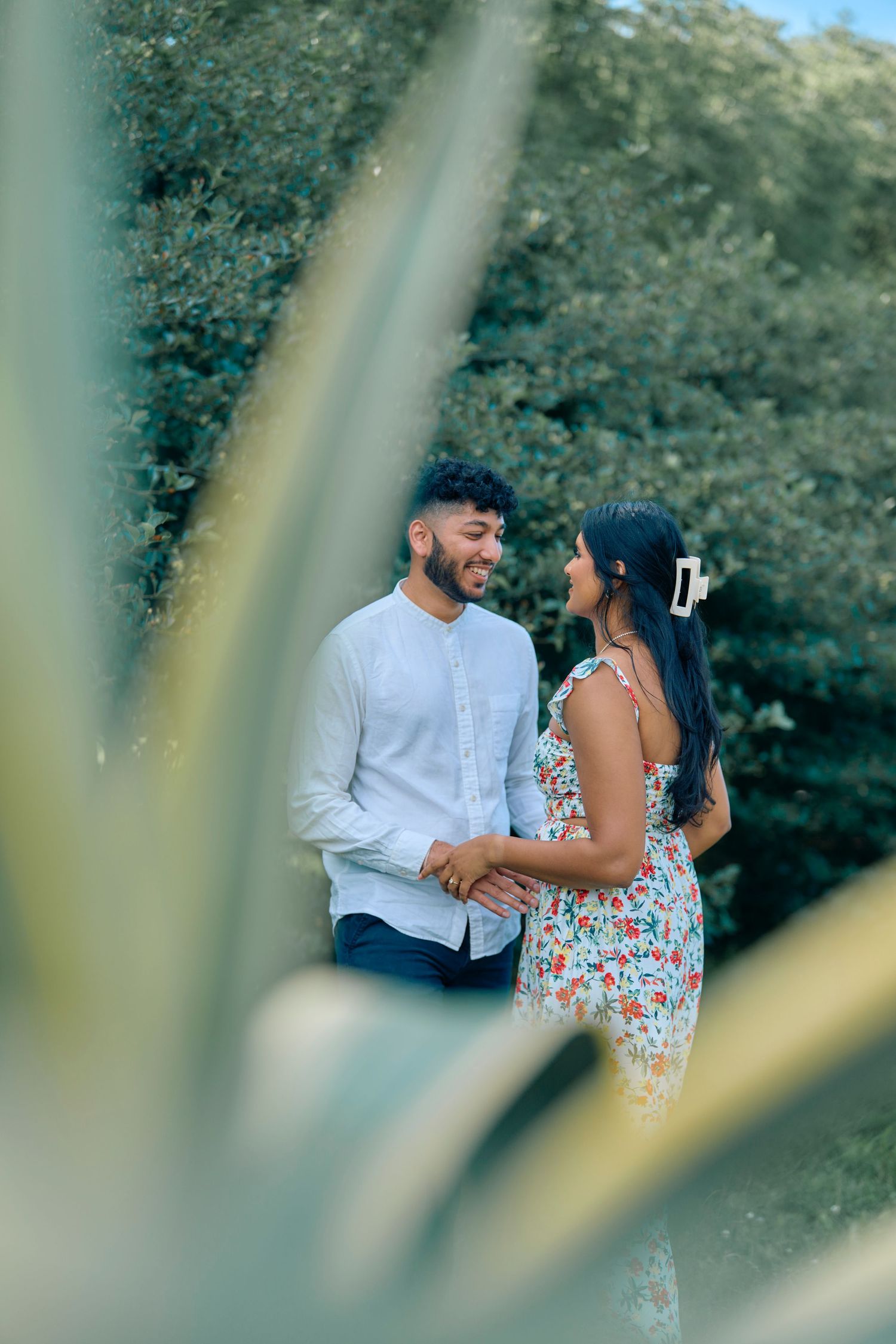 A couple shares a romantic moment in nature, photographed through leaves in the foreground creating a dreamy effect.