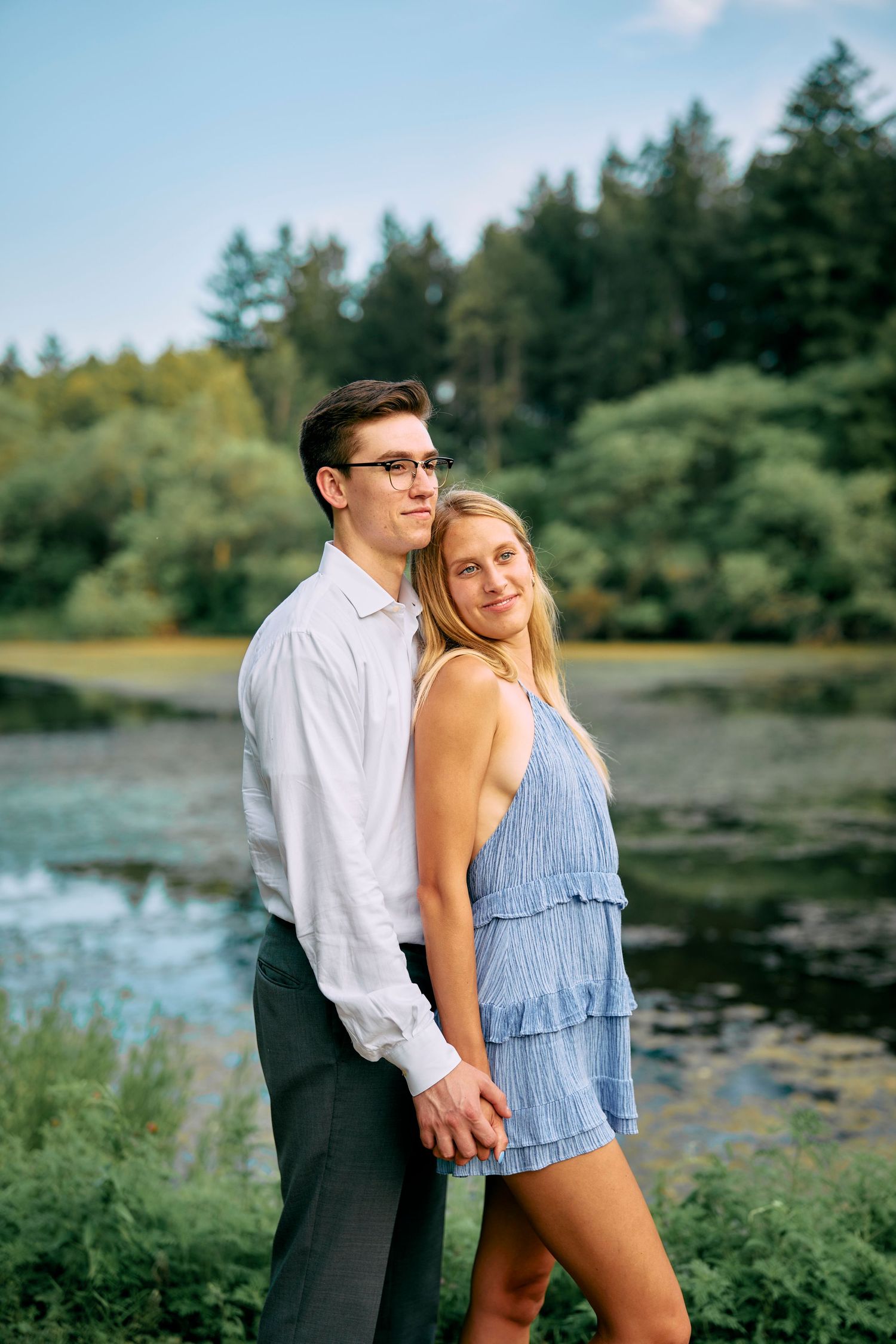 A couple stands together by a scenic waterfront with evergreen trees in the background on a sunny summer day.