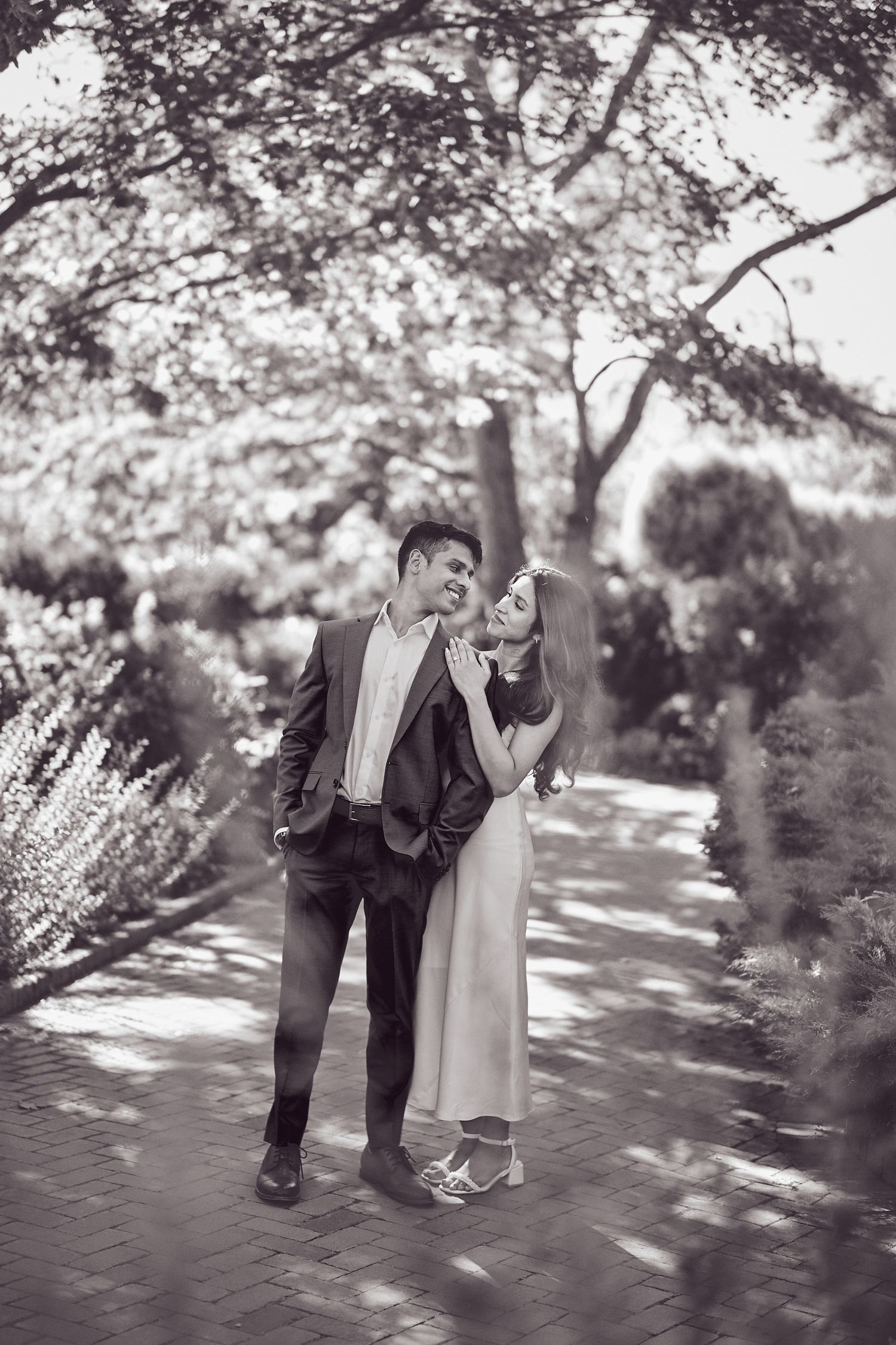 Two people in elegant attire share a joyful moment together on a tree-lined path dappled with sunlight.
