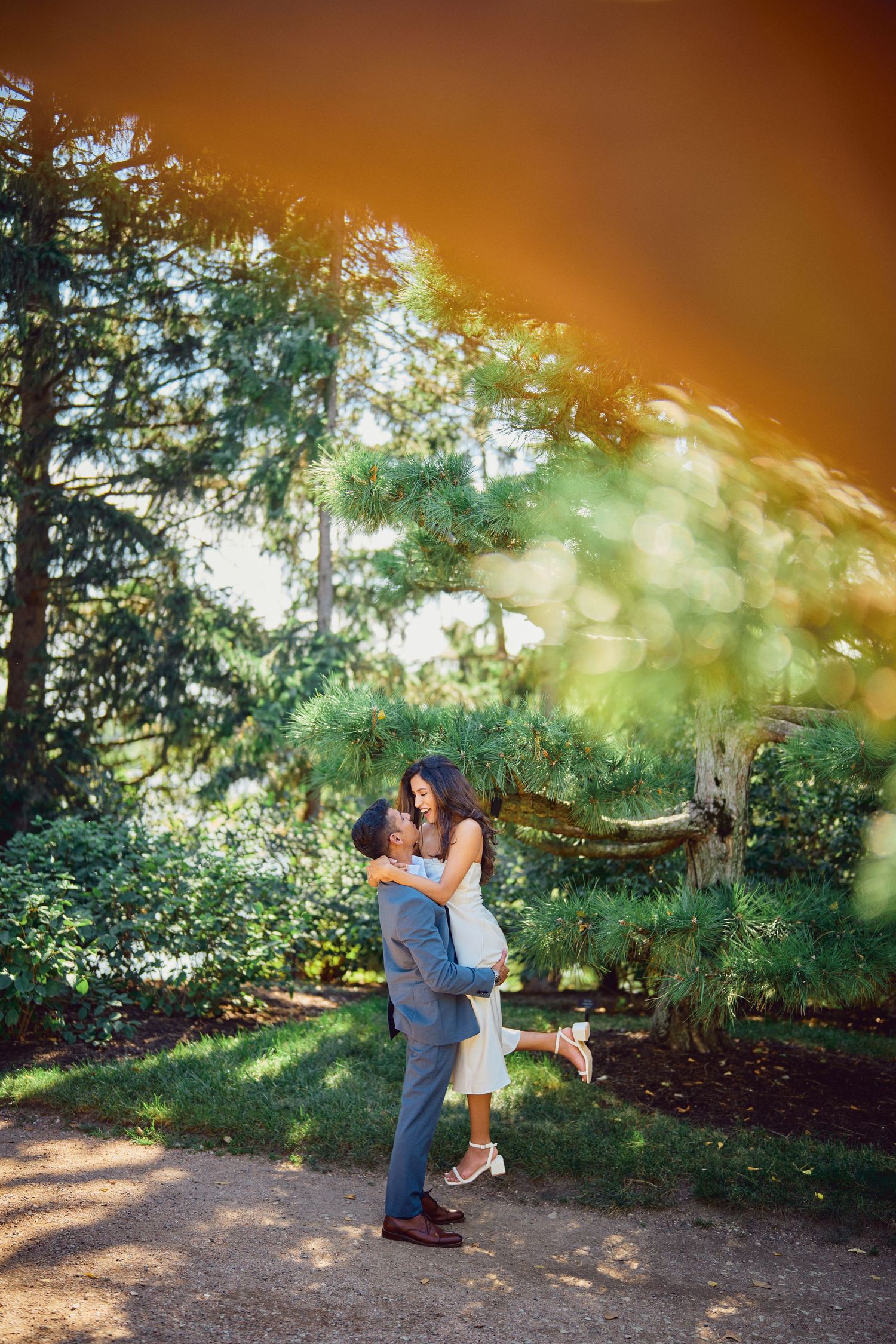 A couple embraces in a romantic outdoor garden setting with sunlit trees and dreamy lighting effects.