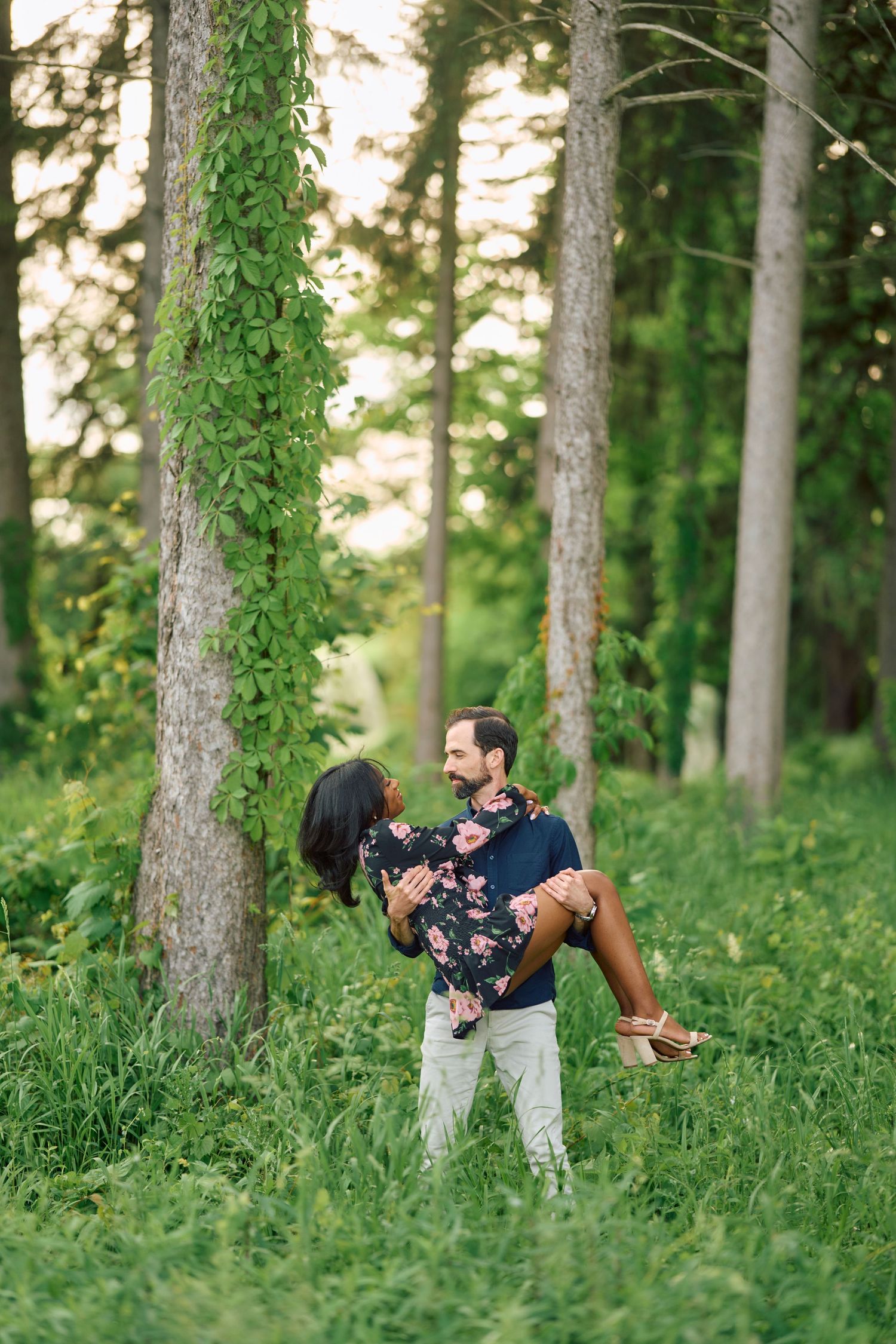 A couple shares a romantic moment in a sun-dappled forest setting with lush greenery and tall trees around them.