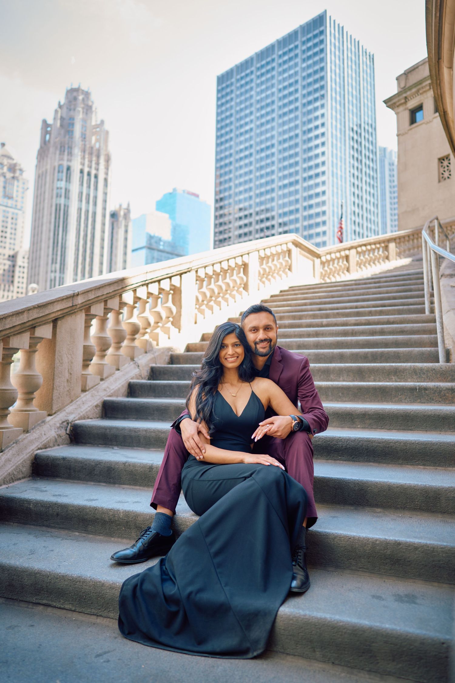 A couple in formal attire poses on elegant stone steps with Chicago skyscrapers rising in the background.