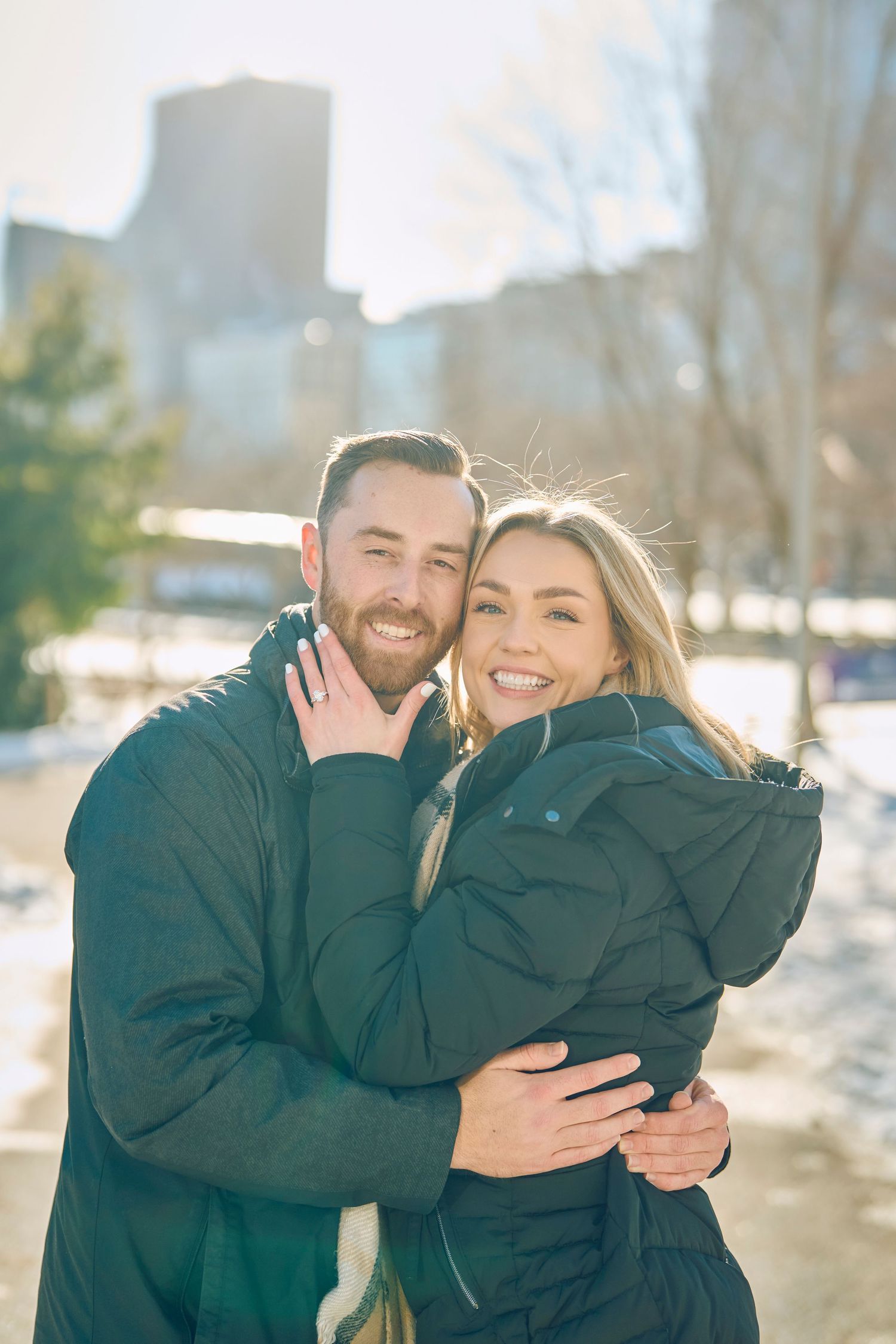 A happy couple embraces outdoors on a sunny winter day with snowy backdrop and city buildings in the distance.