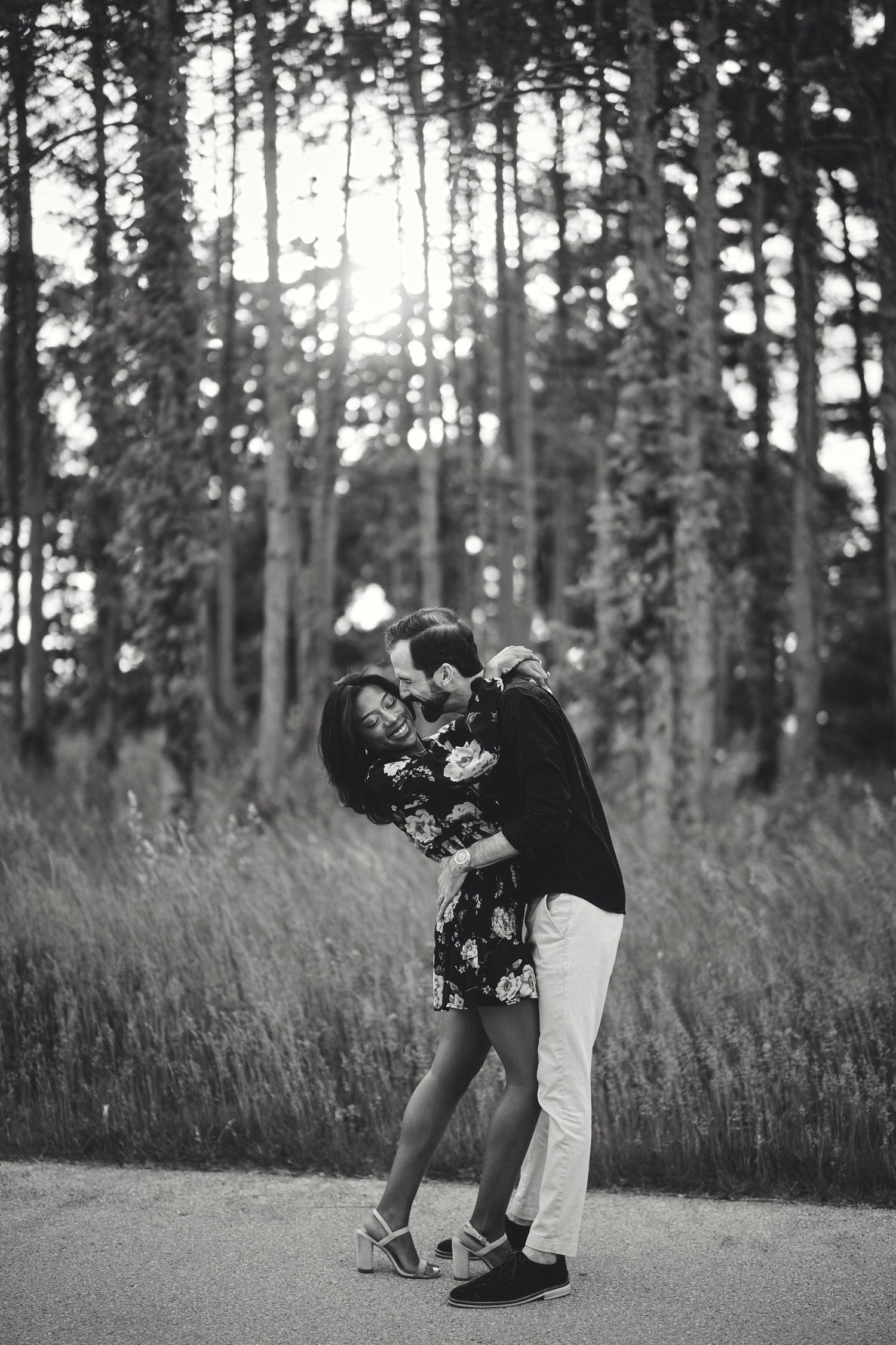 A romantic black and white photo of a couple embracing on a forest path with sunlight filtering through tall trees.