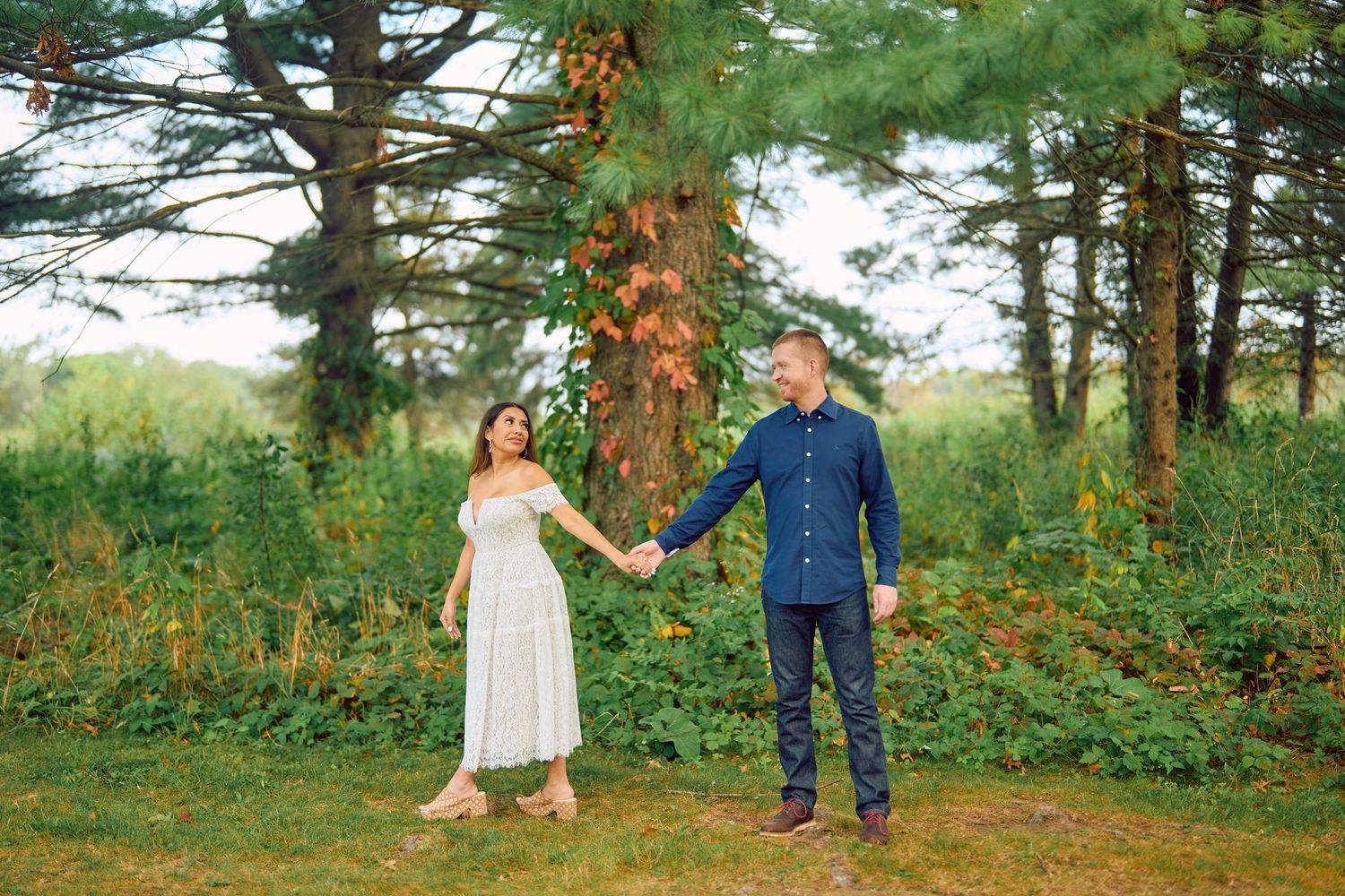 A couple holds hands while standing among tall pine trees in a scenic outdoor forest setting.