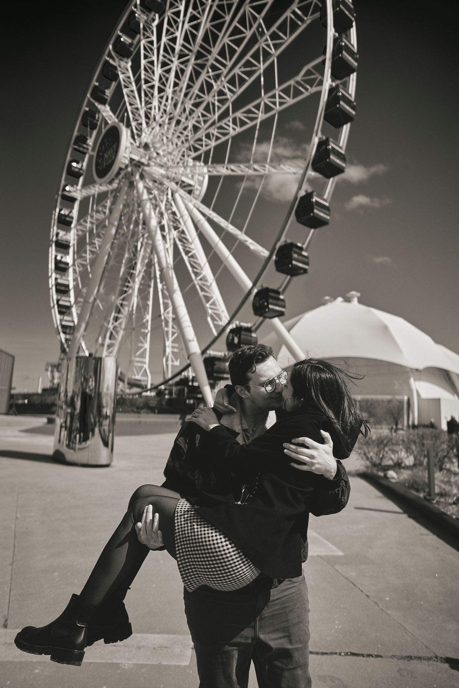 A romantic couple embracing near a large Ferris wheel in black and white photography.