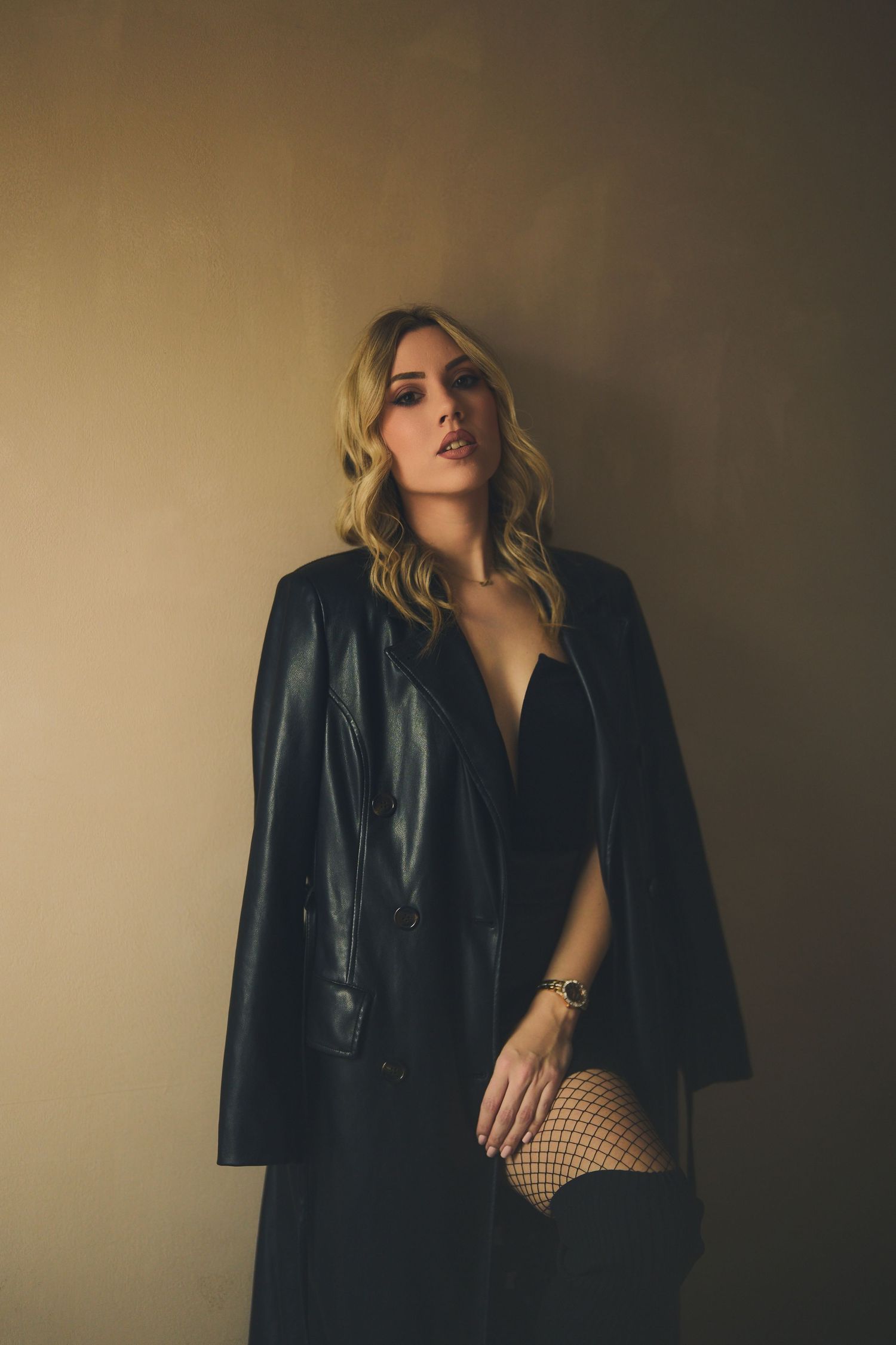 Moody fashion portrait of someone in a black leather jacket and fishnet stockings posing against dark wall.