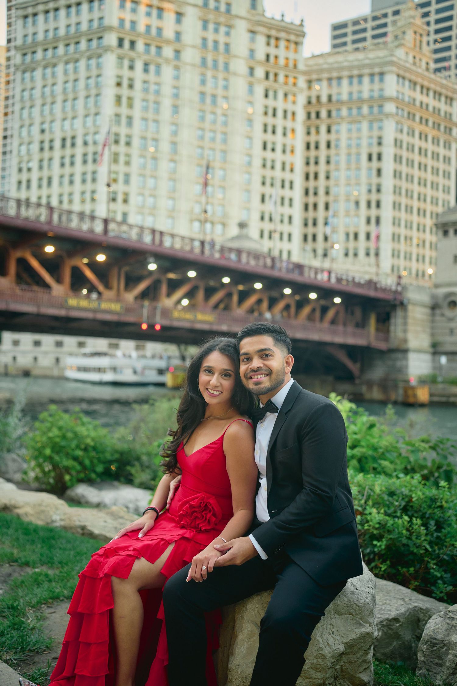 A couple in formal attire poses on rocks near a bridge in downtown Chicago with skyscrapers in the background.