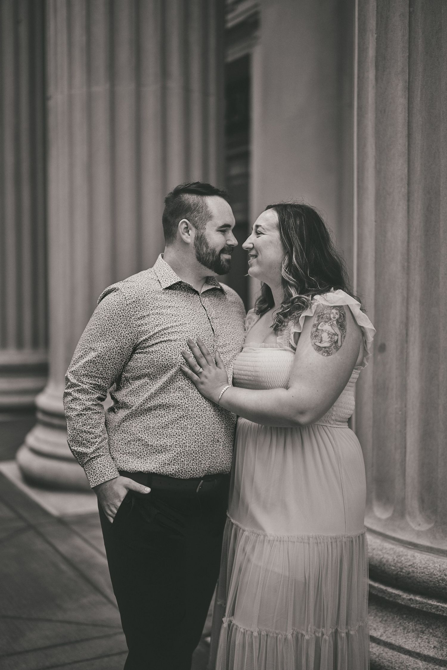 A couple shares an intimate moment in front of classical columns, with the person in a flowing blush dress and visible tattoos.