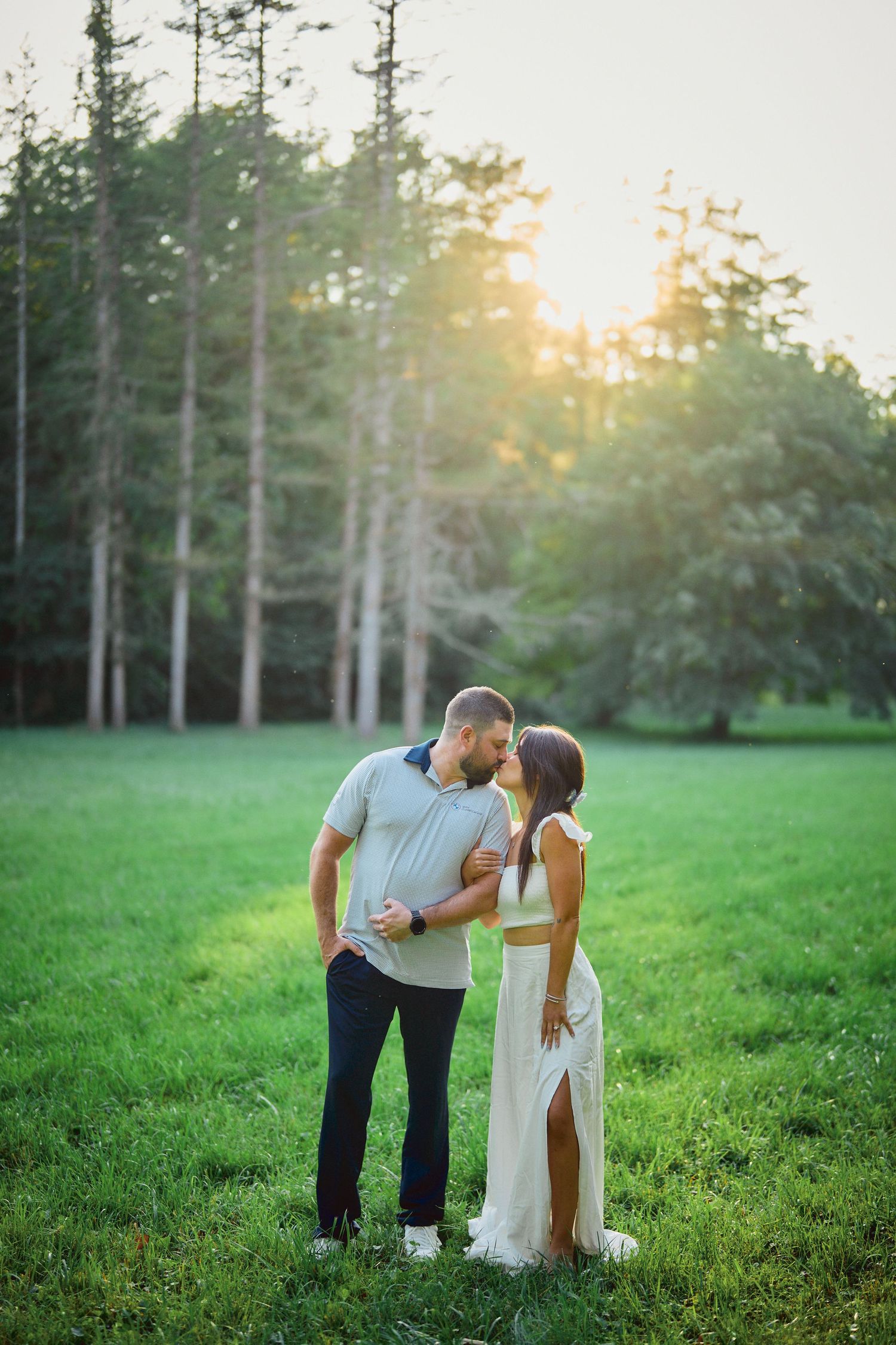 A couple shares an intimate moment in a sun-dappled field with tall evergreen trees in the background.