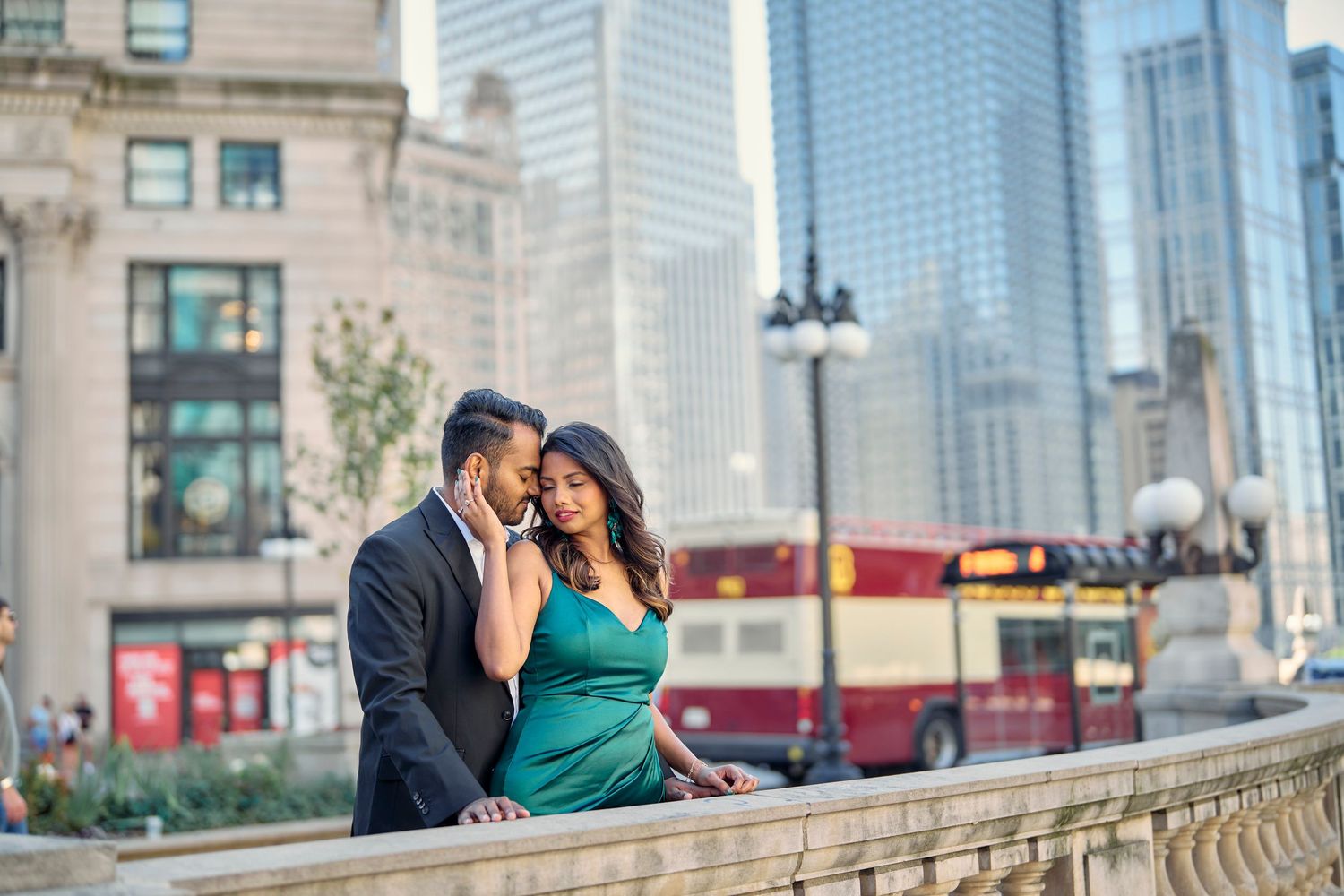 A couple shares an intimate moment along a city street with modern skyscrapers and a red bus in the background.