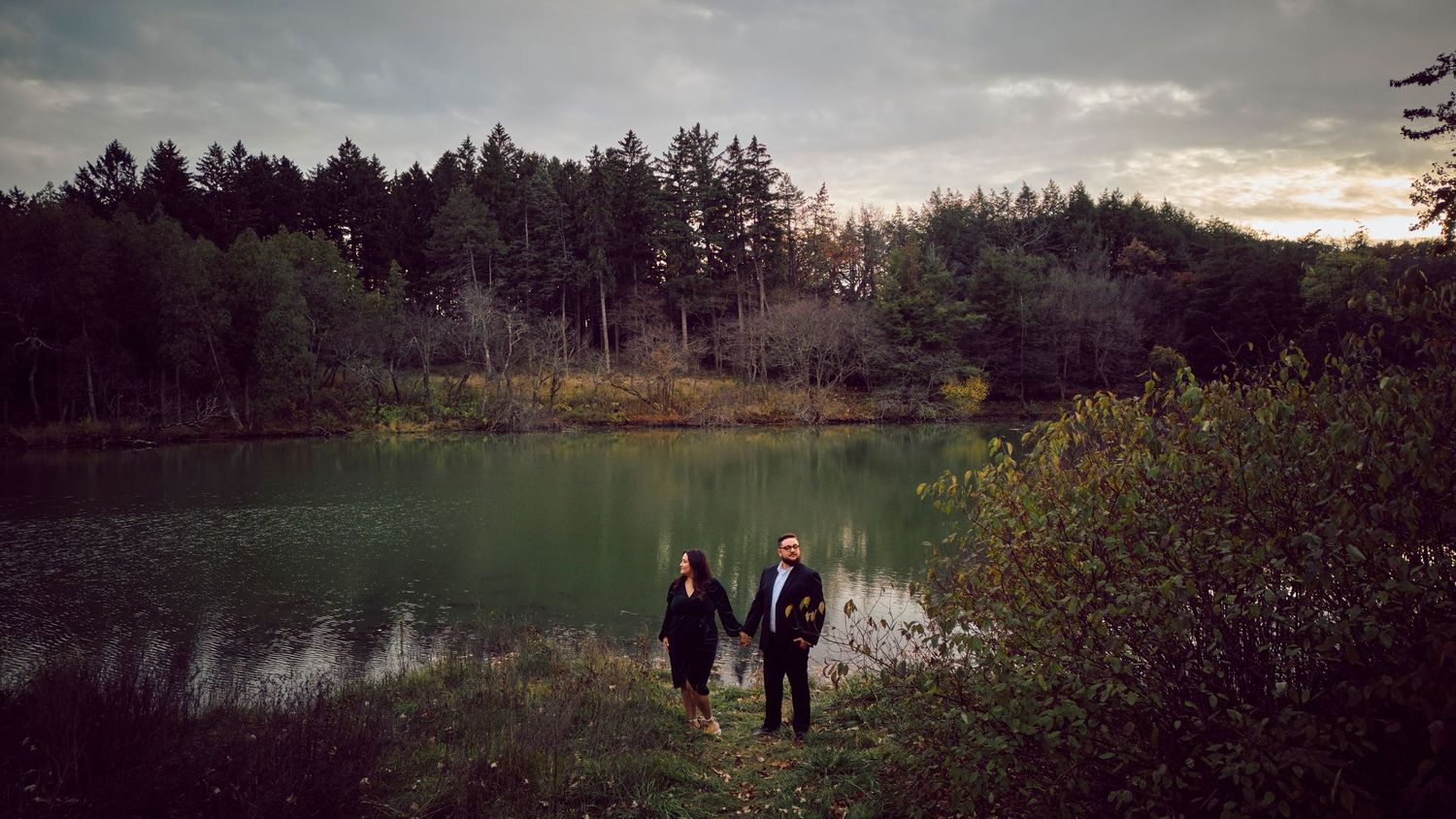 People standing by a tranquil green lake surrounded by pine trees at dusk with moody overcast sky.