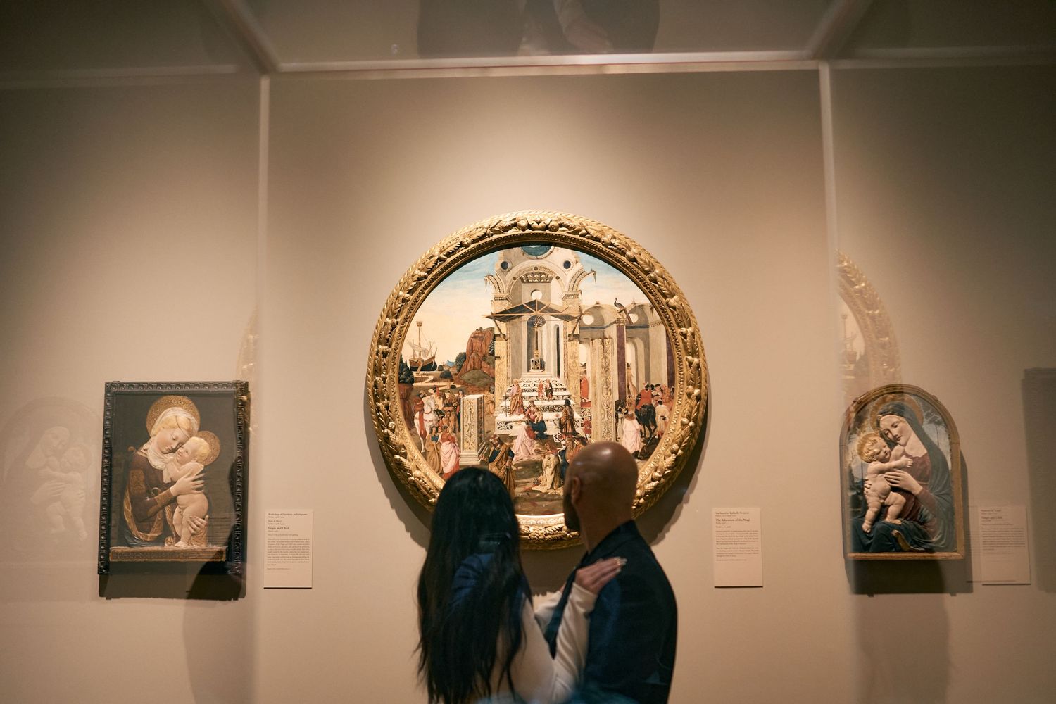 Two visitors view a circular framed religious artwork flanked by smaller paintings in an art gallery setting.