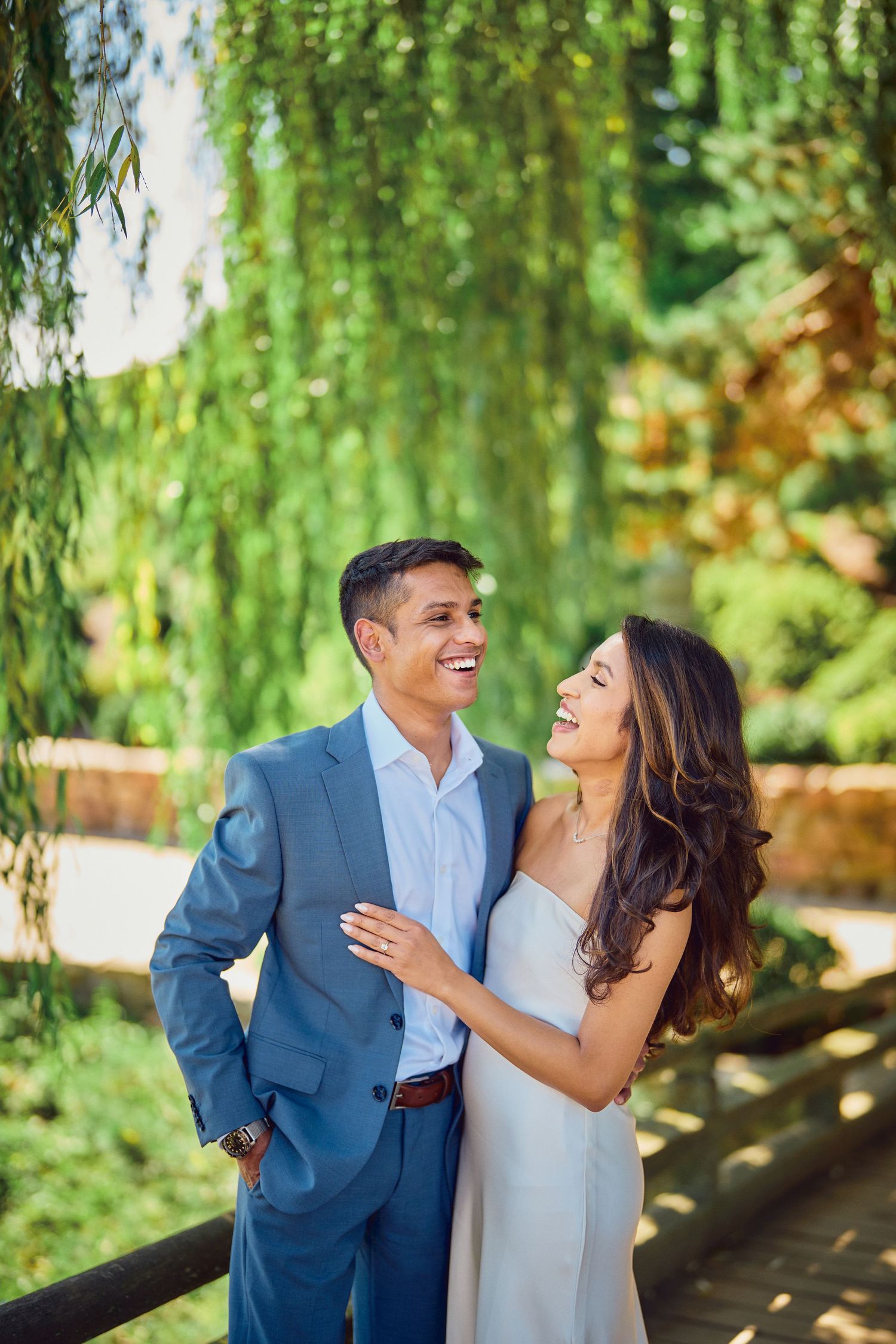A joyful couple shares a candid moment together while standing under a weeping willow tree in a garden setting.