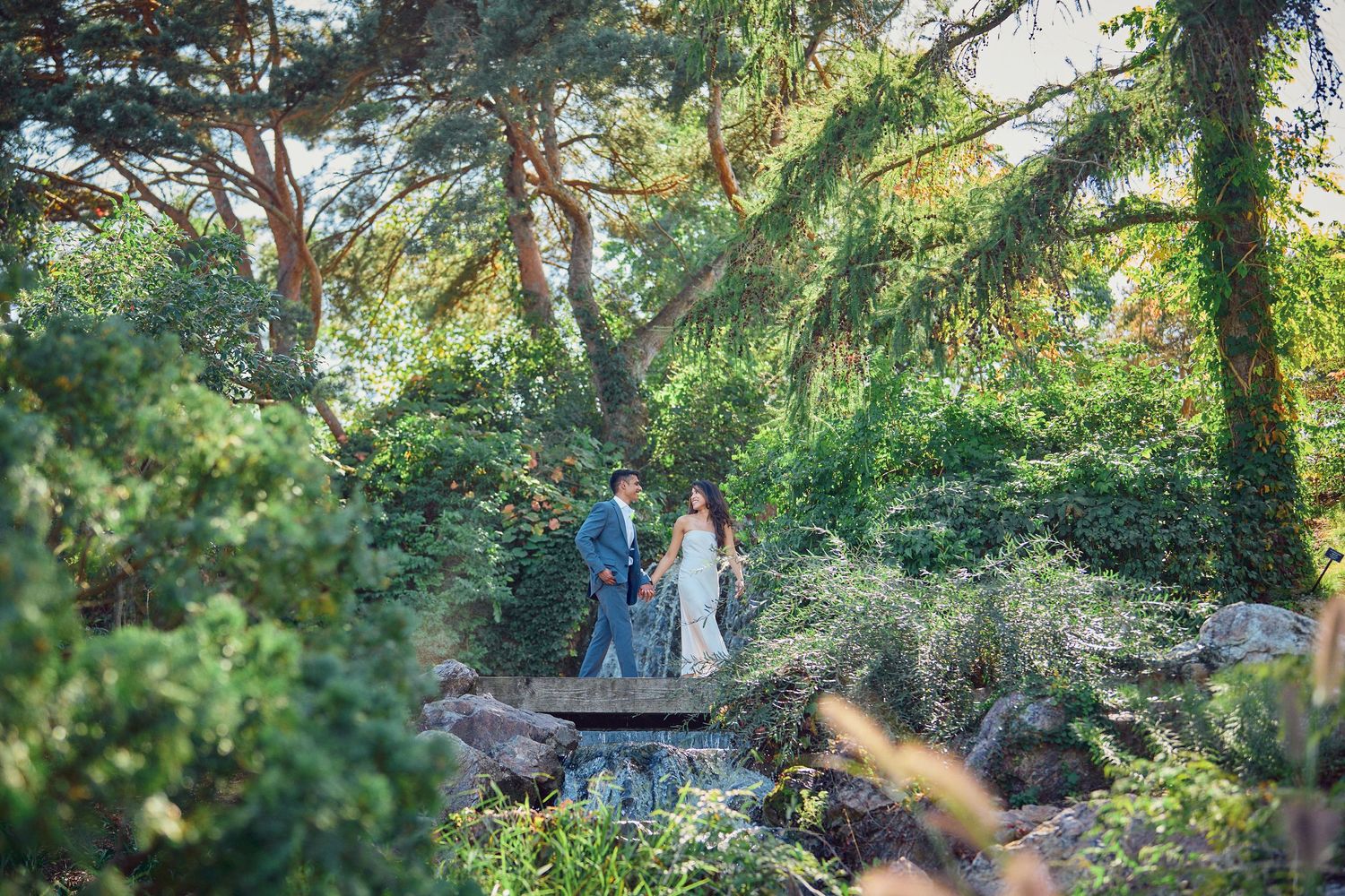 Two people share a moment on a garden path beneath towering pine trees and greenery in an outdoor setting.