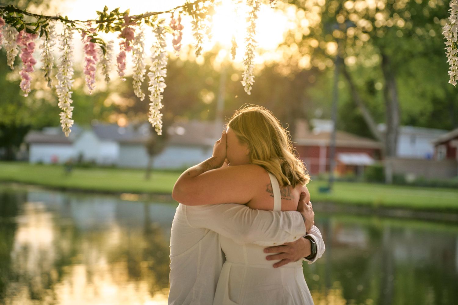A warm embrace between two people under hanging wisteria flowers at sunset by a tranquil pond.