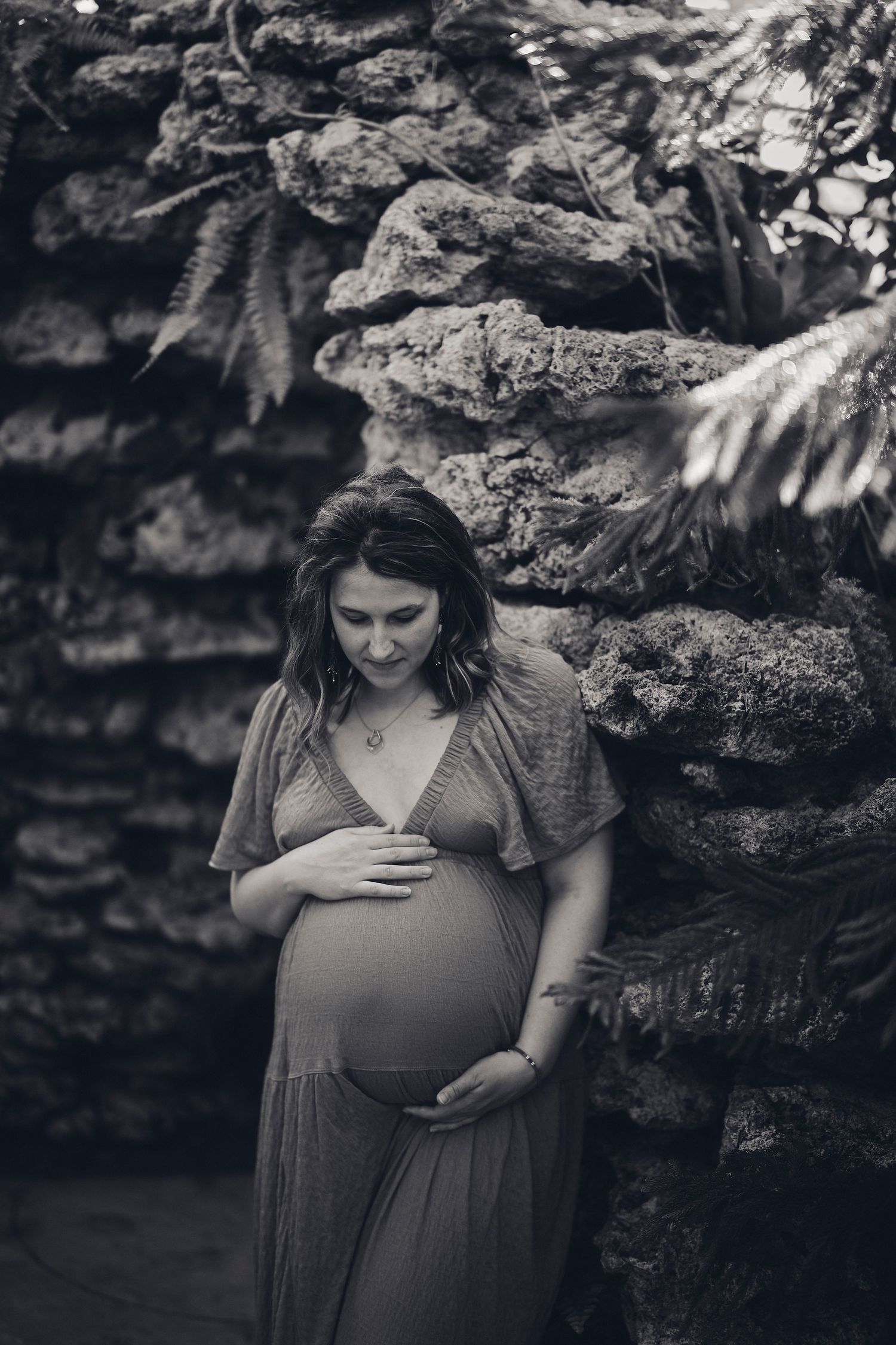 Black and white maternity photo showing an expectant mother in a flowing dress against a stone wall backdrop.