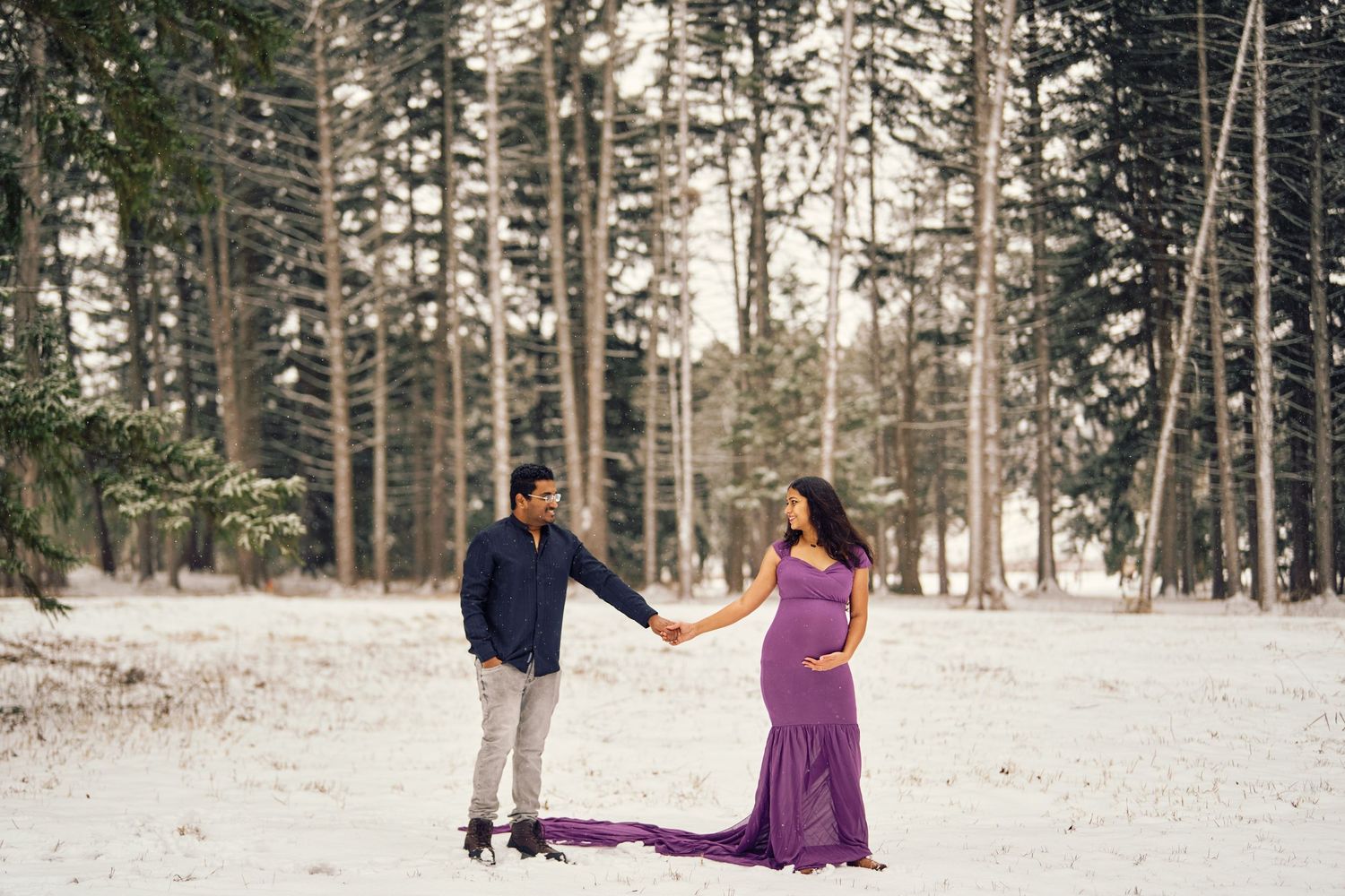 A couple stands holding hands in a snowy forest setting with a flowing purple dress catching the winter light.