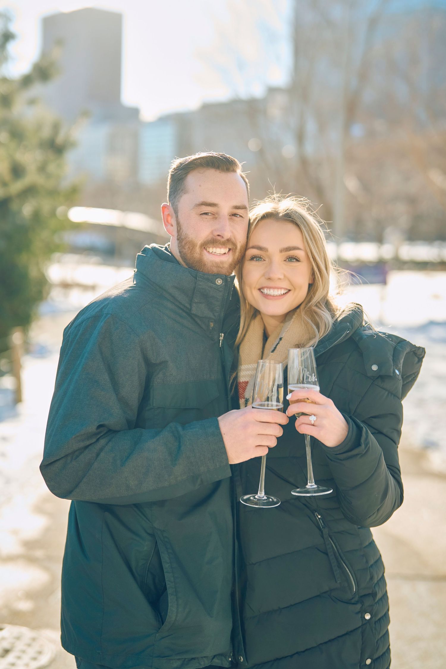 A couple in winter coats toasts with champagne glasses while smiling outdoors on a sunny winter day in a snowy setting.