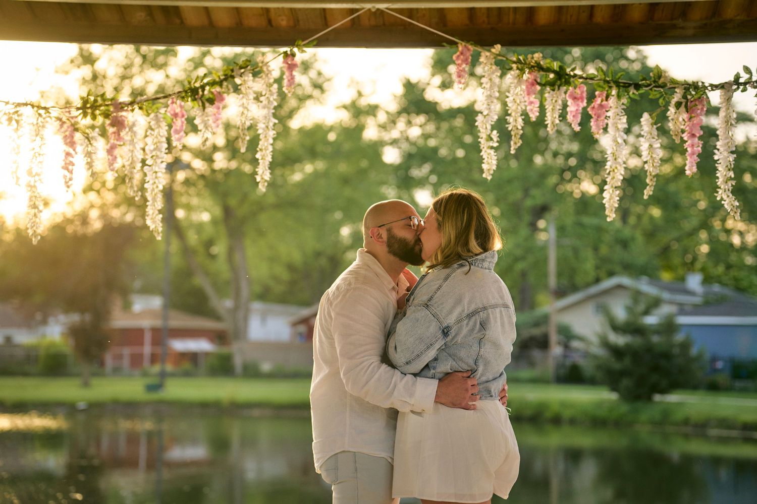 Two silhouettes embrace under hanging floral decorations by a lake during golden hour sunset.