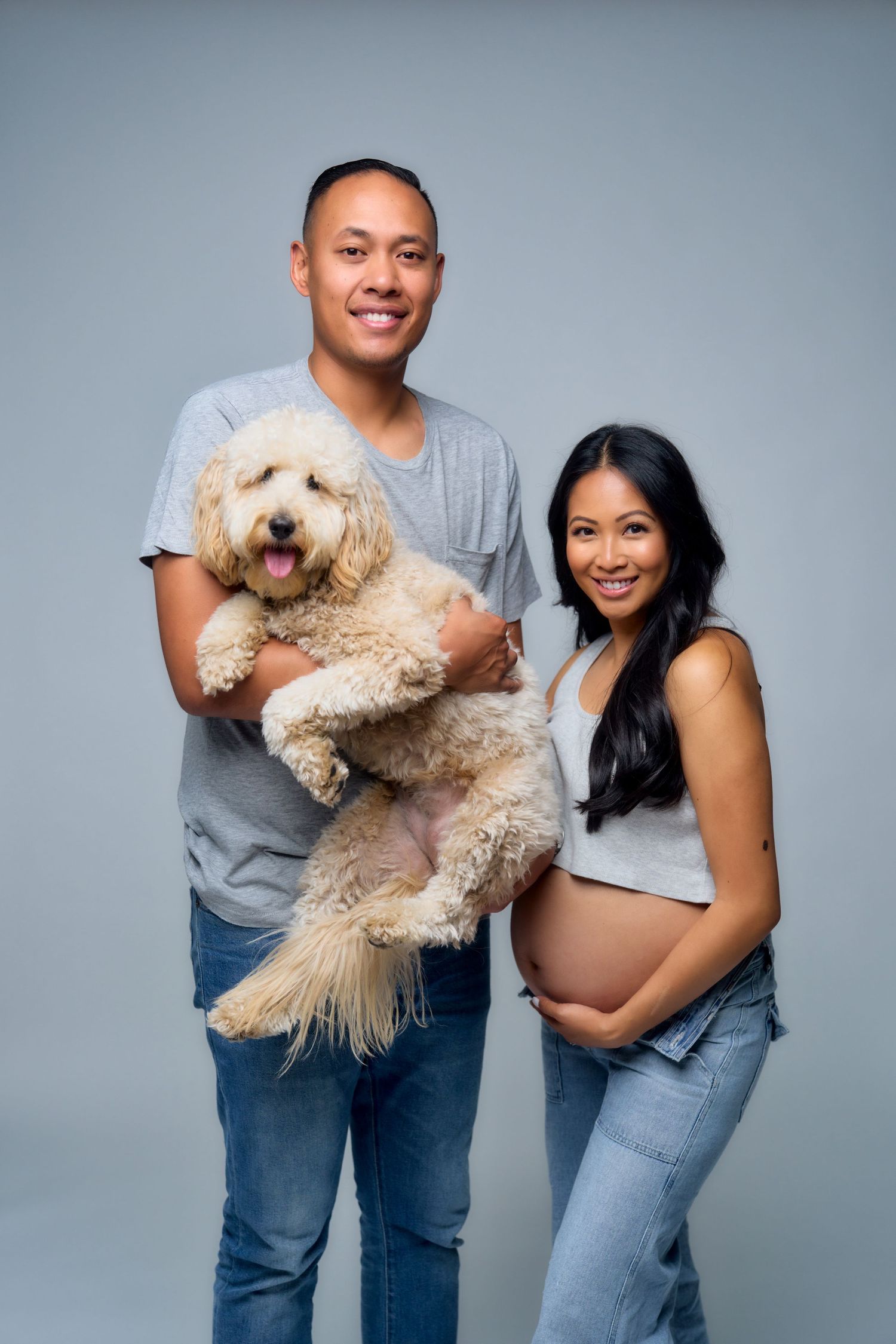 A couple poses with their fluffy goldendoodle dog while expecting a baby in a studio portrait against a gray background.
