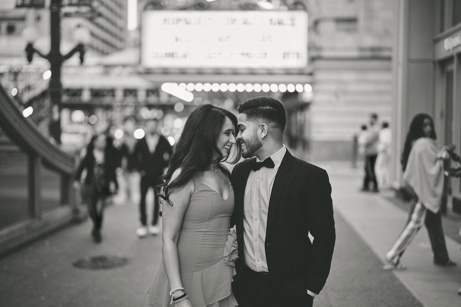 Romantic couple in formal attire share an intimate moment on a busy city street in black and white photography.